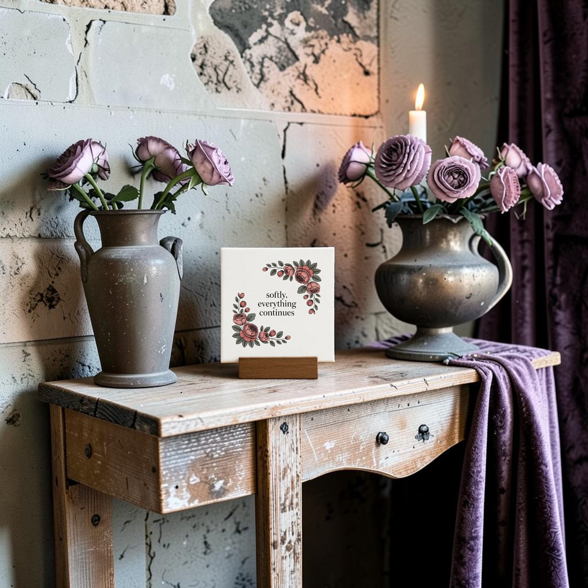 A rustic wooden table displays two vases with purple roses, a lit candle, and the Mirabilia Boutique Softly, Everything Continues Mini Sign—a decorative piece—against a textured stone wall and draped purple fabric.