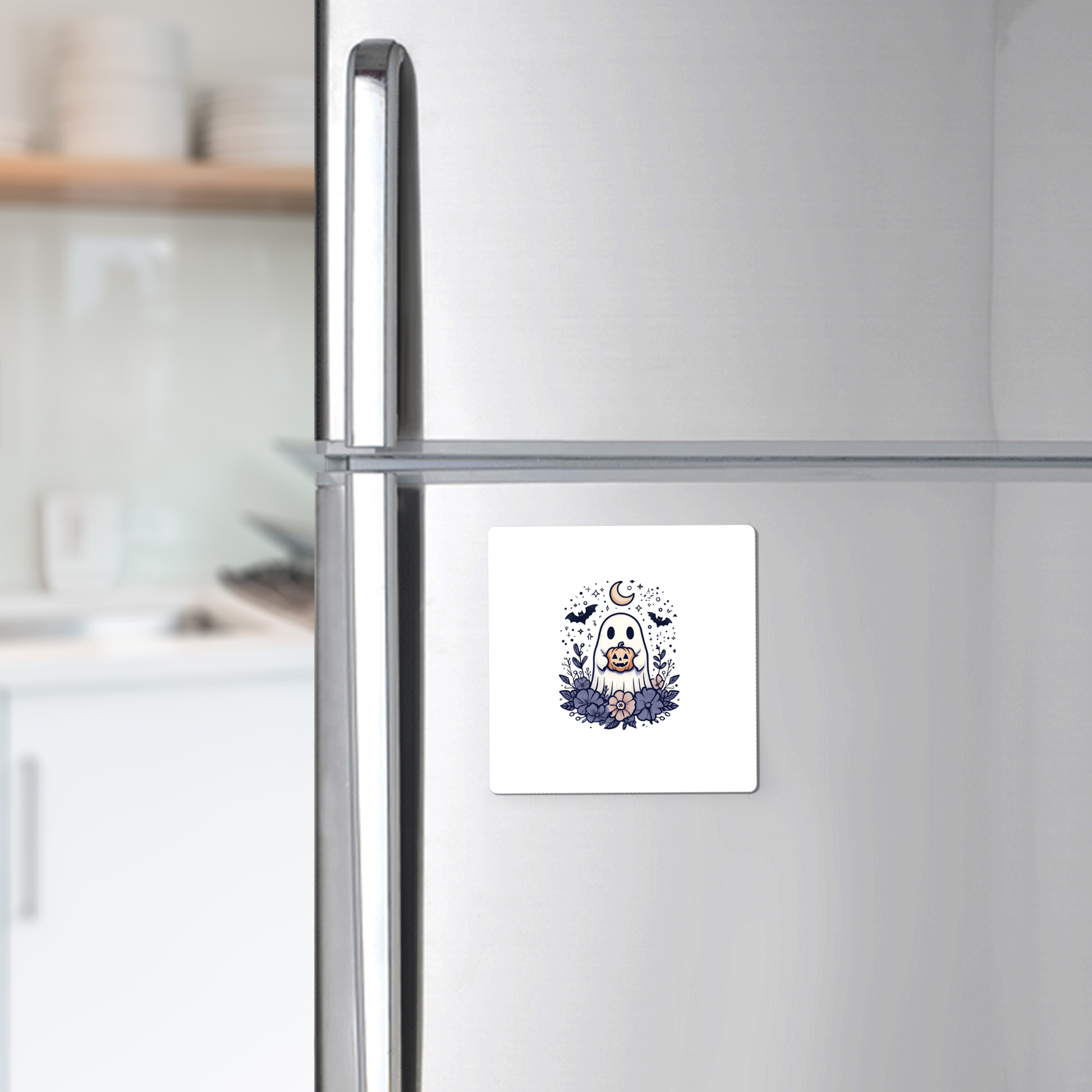 A silver refrigerator features a square white magnet displaying an adorable owl, moon, and stars illustration, bringing charm to your kitchen. For a festive touch this season, pair it with the Cute Ghost Pumpkin Magnet from Mirabilia Boutique—its whimsical Halloween design complements your other decorative accents beautifully. Kitchen shelves and dishes can be seen in the background.