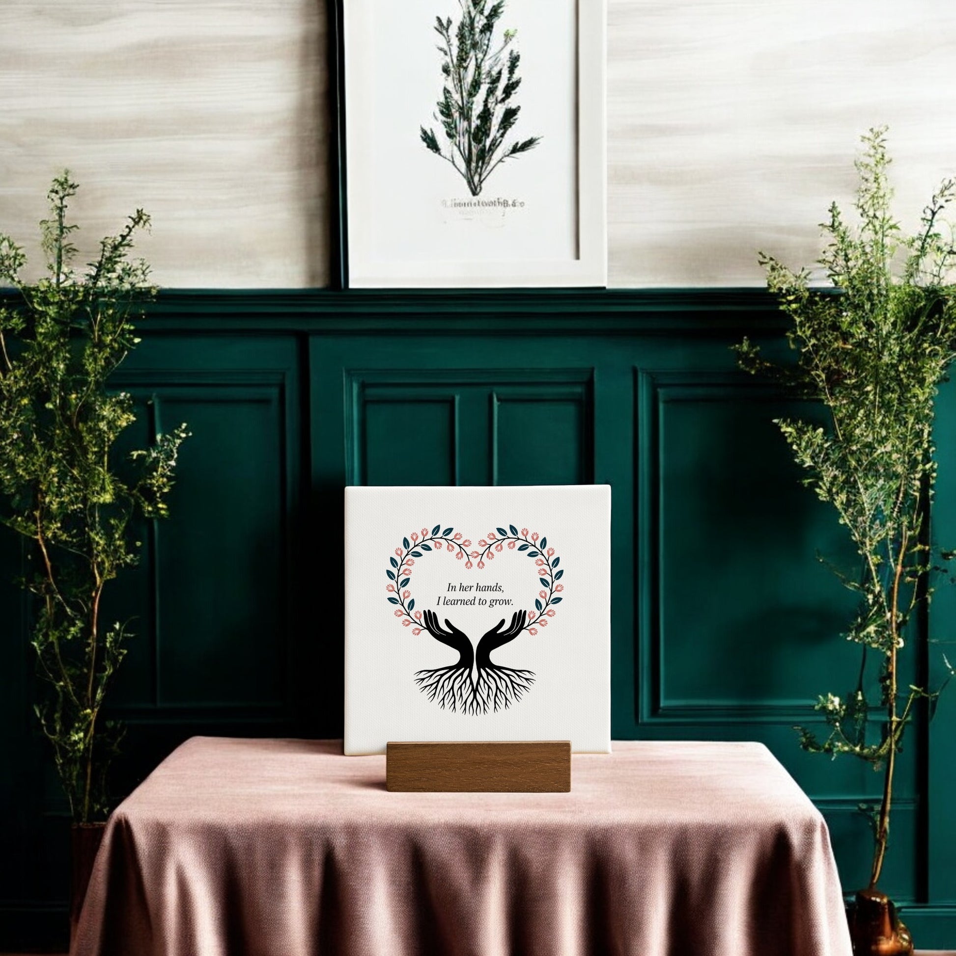The In Her Hands Mini Sign by Mirabilia Boutique features a tree with heart-shaped branches and the words, “In her hands, I learned to grow”—a thoughtful Mother’s Day gift displayed on a mauve cloth amid greenery and art.