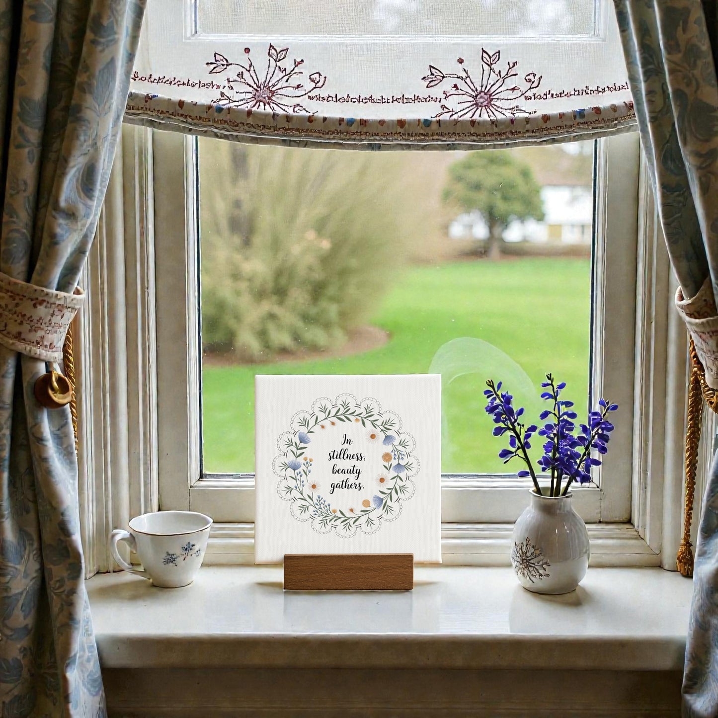 A windowsill with a cup, purple flowers, and the "In Stillness Beauty Gathers Mini Canvas Sign" by Mirabilia Boutique, paired with an embroidered curtain and garden view—ideal as meditation shelf decor.