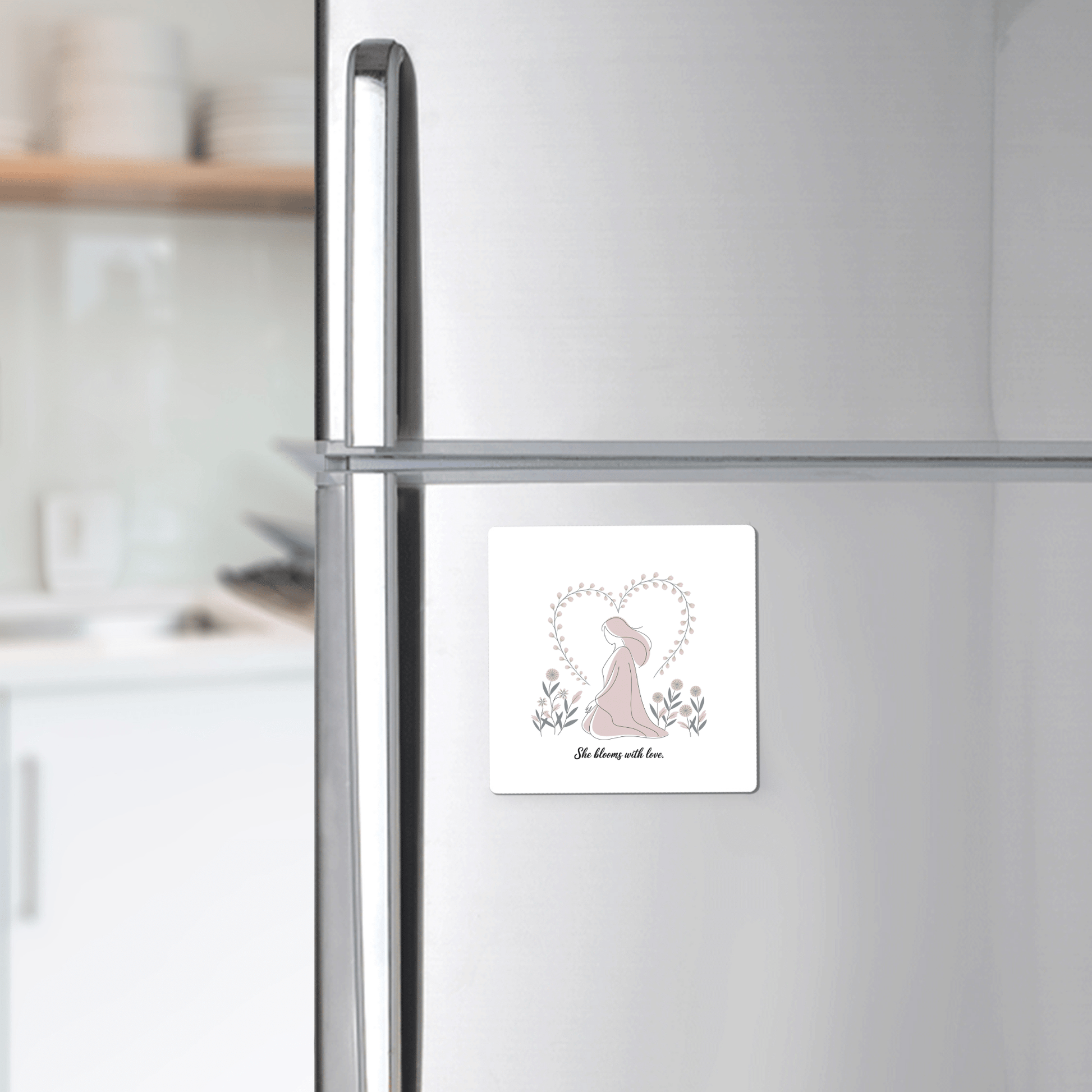 A silver refrigerator features the She Blooms with Love Magnet – Gentle Floral Mom Gift by Mirabilia Boutique, a square décor piece with a bunny and leafy heart—perfect for celebrating motherhood. The bright kitchen creates a warm, inviting backdrop.
