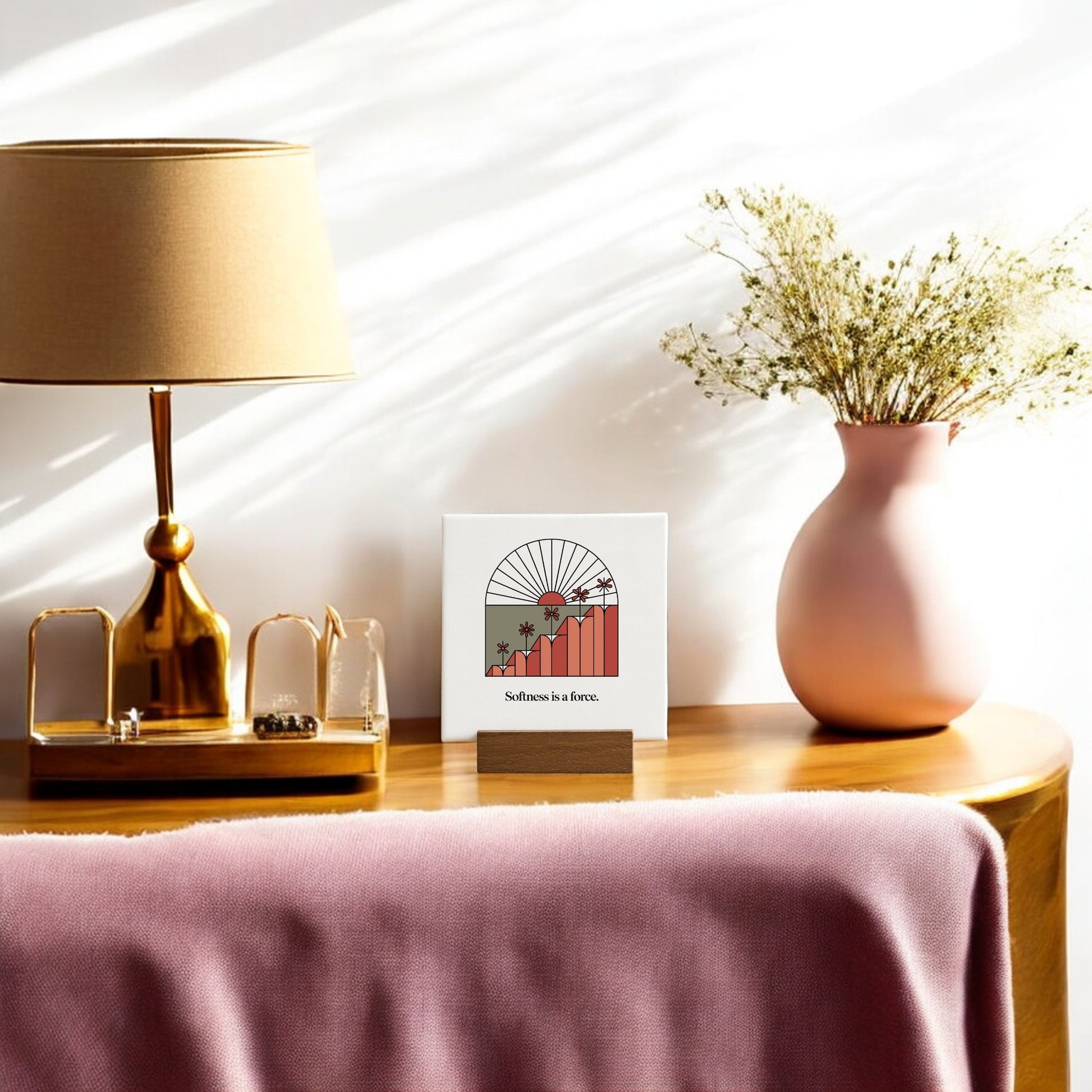 A Softness Is a Force Mini Canvas Sign by Mirabilia Boutique sits on a wooden console table with a gold lamp, pink vase of dried flowers, and pink throw blanket—perfect for showcasing minimalist or motivational desk decor.