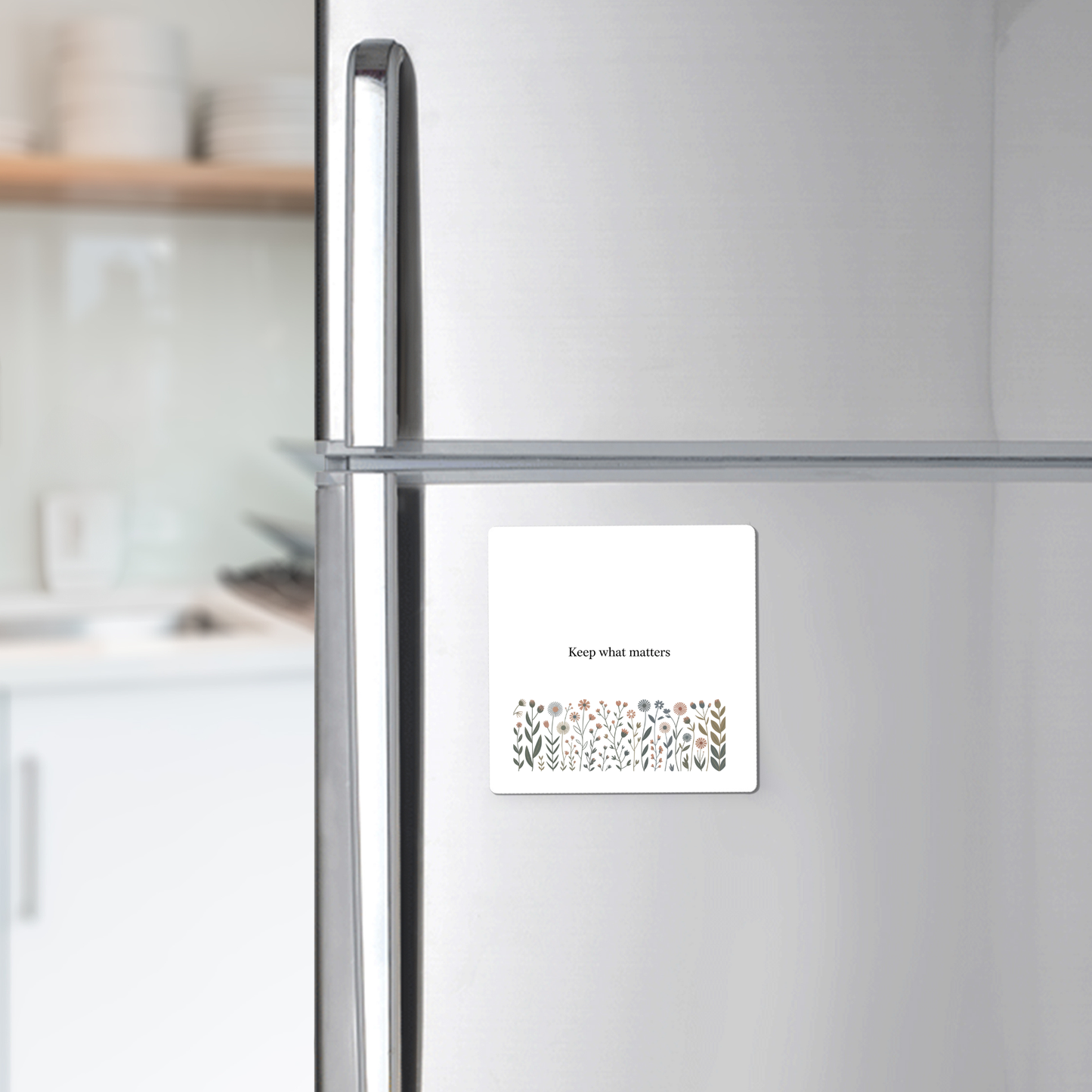 A Mirabilia Boutique Keep What Matters Floral Magnet, featuring an inspiring quote and flower illustration, adds a charming touch to any stainless steel fridge—ideal for gifting or enhancing cottage décor. A blurred kitchen is seen in the background.