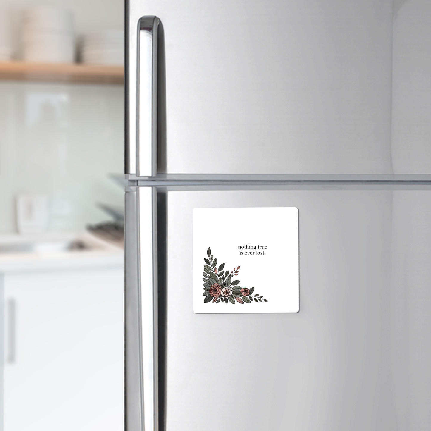 A silver refrigerator holds the Mirabilia Boutique "Nothing True Is Ever Lost Floral Magnet," featuring delicate corner artwork. Ideal as an encouragement gift, it stands out with dishes on a kitchen counter in the background.