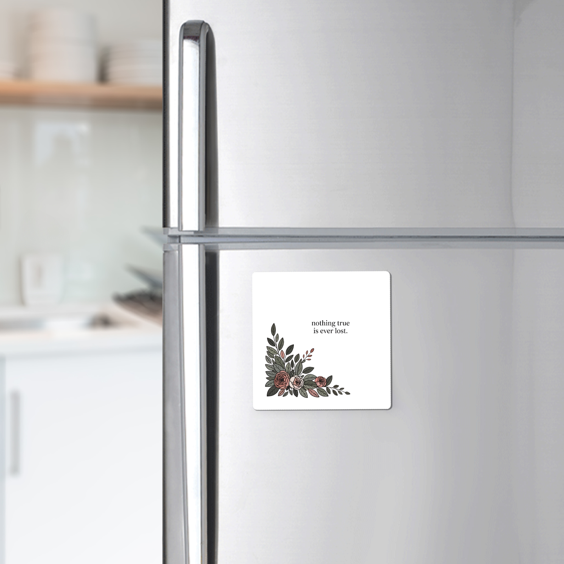 A silver refrigerator holds the Mirabilia Boutique "Nothing True Is Ever Lost Floral Magnet," featuring delicate corner artwork. Ideal as an encouragement gift, it stands out with dishes on a kitchen counter in the background.