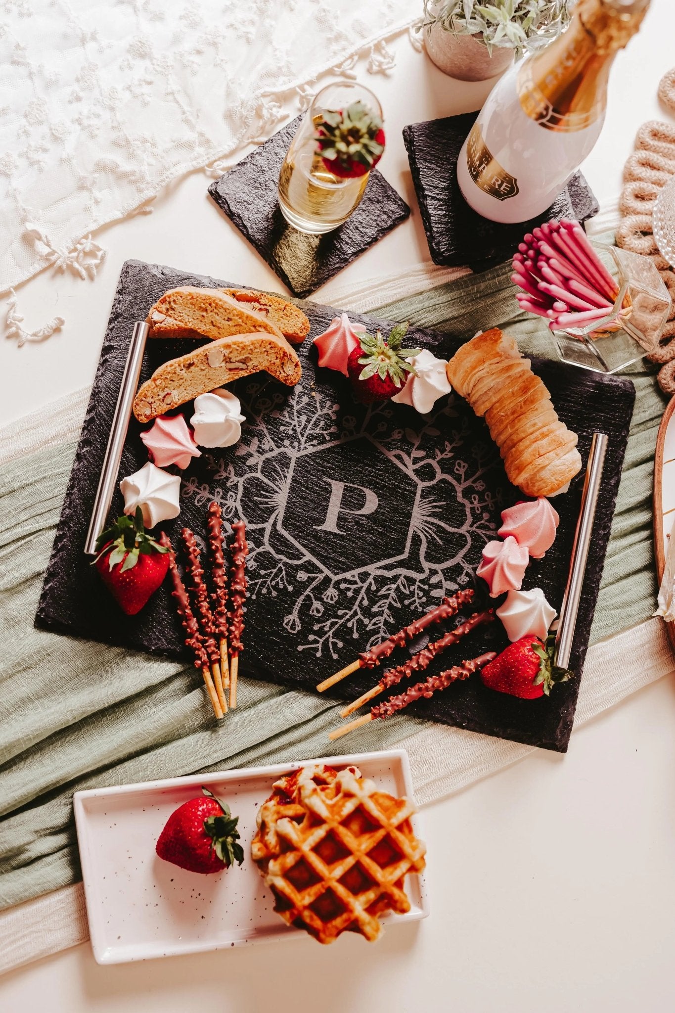 The Mirabilia Boutique Personalized Engraved Slate Serving Tray with Handles, featuring a floral monogram “P”, is elegantly displayed with strawberries, meringues, biscotti, chocolate sticks, a croissant, and a sparkling drink—perfect for gifting or entertaining.