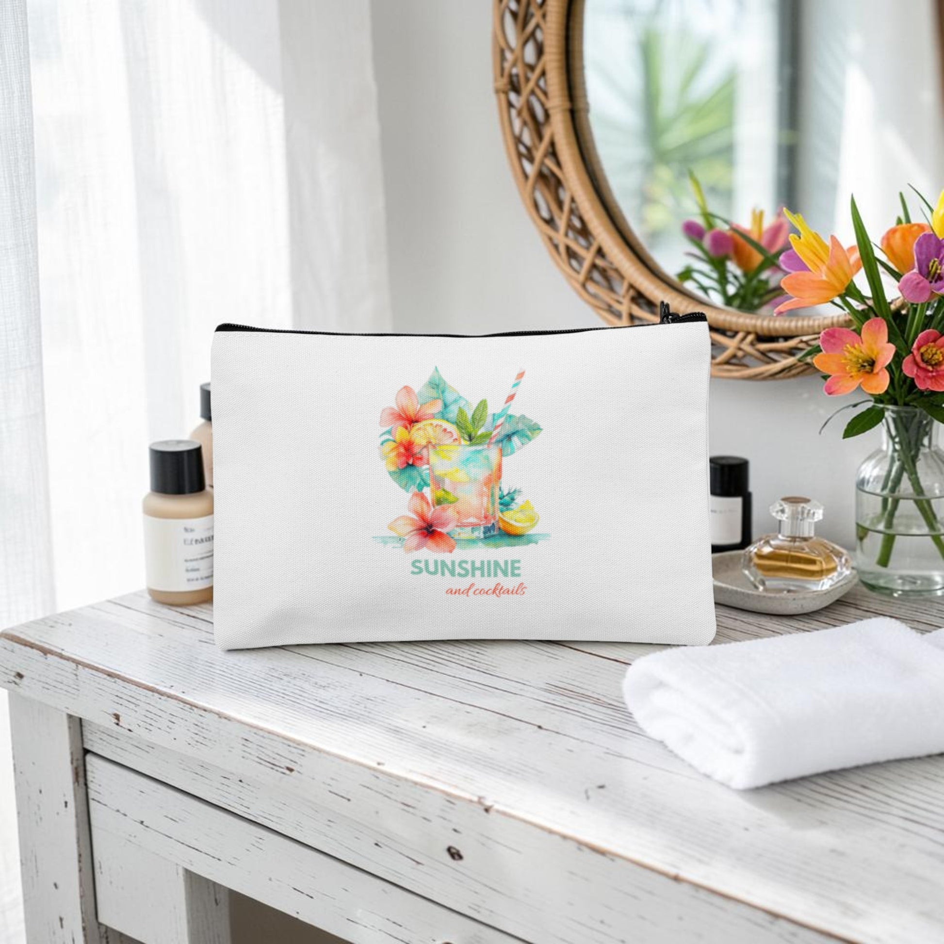 The Sunshine and Cocktails Makeup Bag by Mirabilia Boutique, featuring a vibrant tropical drink and flowers design on white, sits on a wooden vanity near beauty products, a mirror, and a bouquet of fresh flowers.