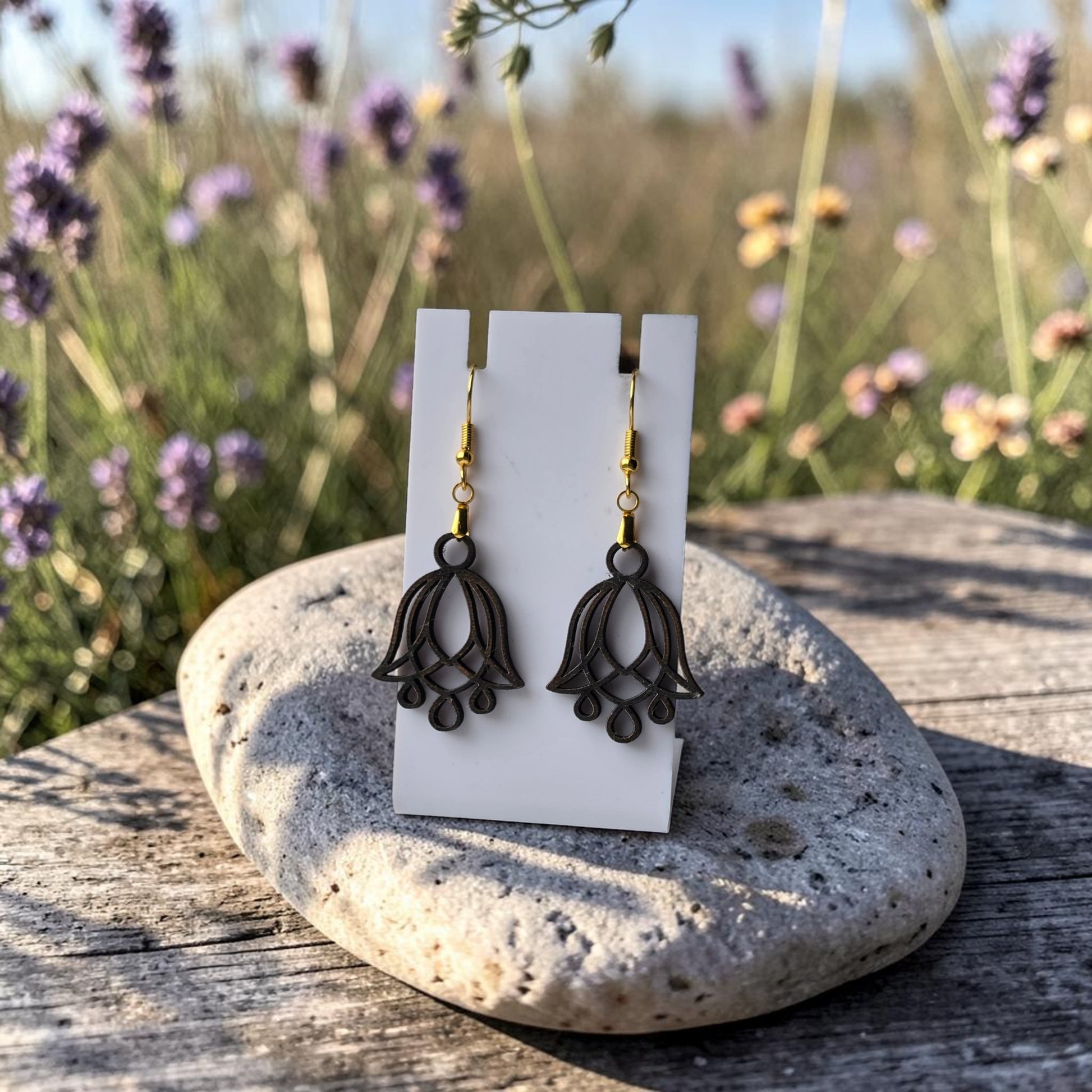 A pair of Bluebell Flower Earrings by Mirabilia Boutique, lasercut from walnut wood with gold hooks, displayed on a white card atop a smooth gray stone outdoors—lavender and wildflowers blurred in the sunny background.