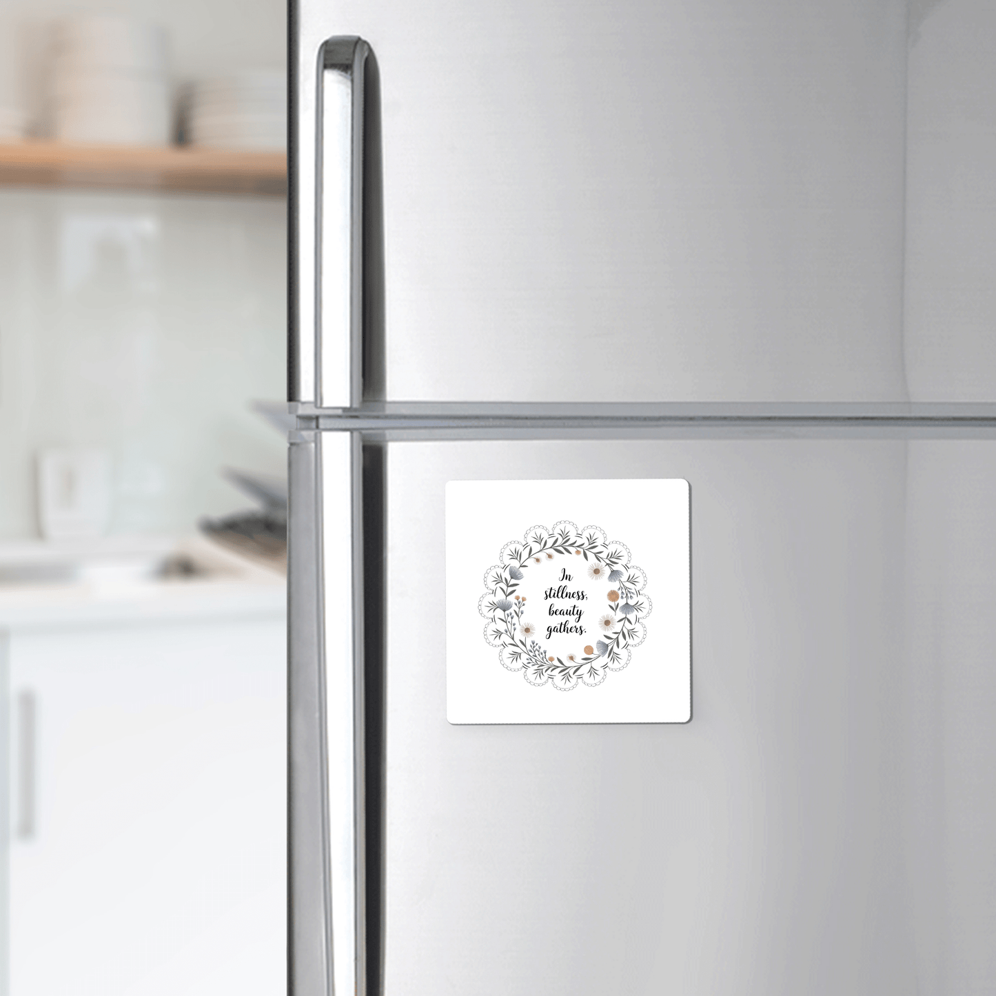 A silver refrigerator displays Mirabilia Boutique's "In Stillness Beauty Gathers" floral quote magnet, featuring a wreath design. A white kitchen with shelves and dishes appears softly blurred in the background, creating mindful decor.