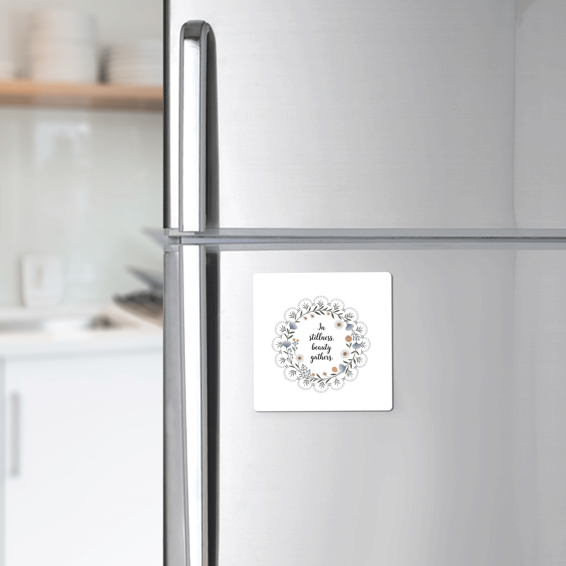 A silver refrigerator displays Mirabilia Boutique's "In Stillness Beauty Gathers" floral quote magnet, featuring a wreath design. A white kitchen with shelves and dishes appears softly blurred in the background, creating mindful decor.