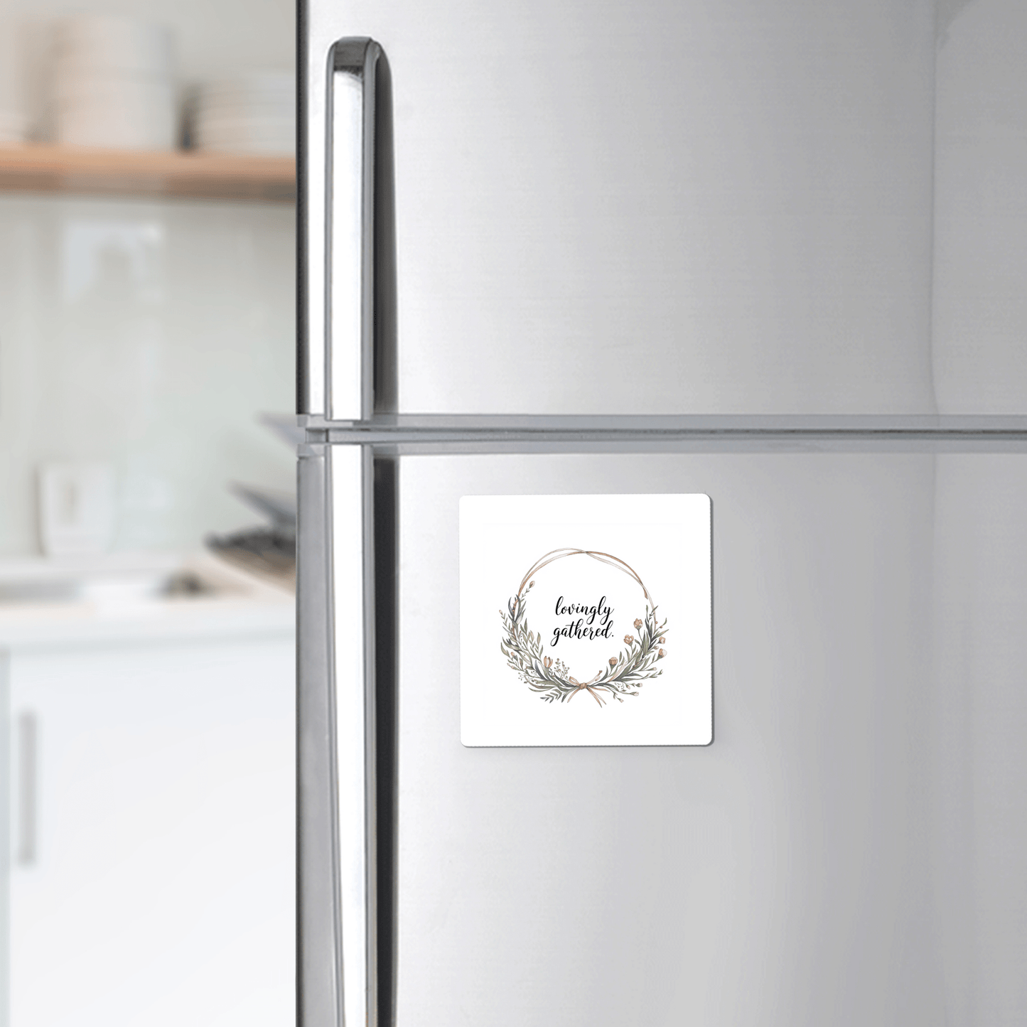 The Mirabilia Boutique "Lovingly Gathered" Magnet, featuring a floral wreath quote design, is shown on a silver fridge in a modern kitchen.