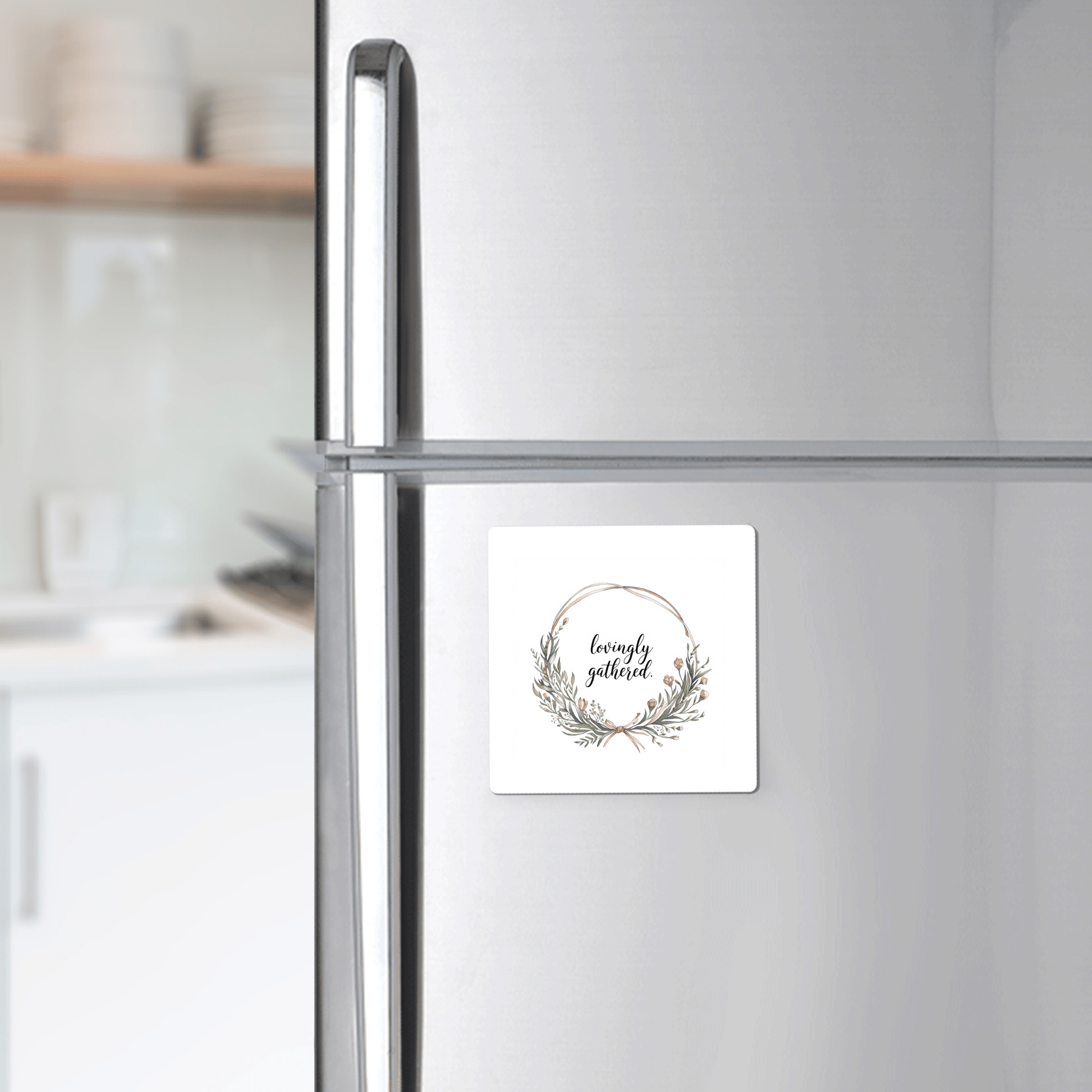 The Mirabilia Boutique "Lovingly Gathered" Magnet, featuring a floral wreath quote design, is shown on a silver fridge in a modern kitchen.