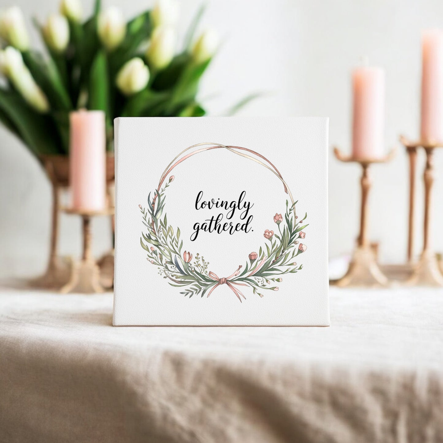 The Mirabilia Boutique Lovingly Gathered Canvas – 8x8 Botanical Wreath Wall Art sits on a beige tablecloth, with white tulips in a vase and three lit pink candles—creating a soft, elegant gathering home decor setting.