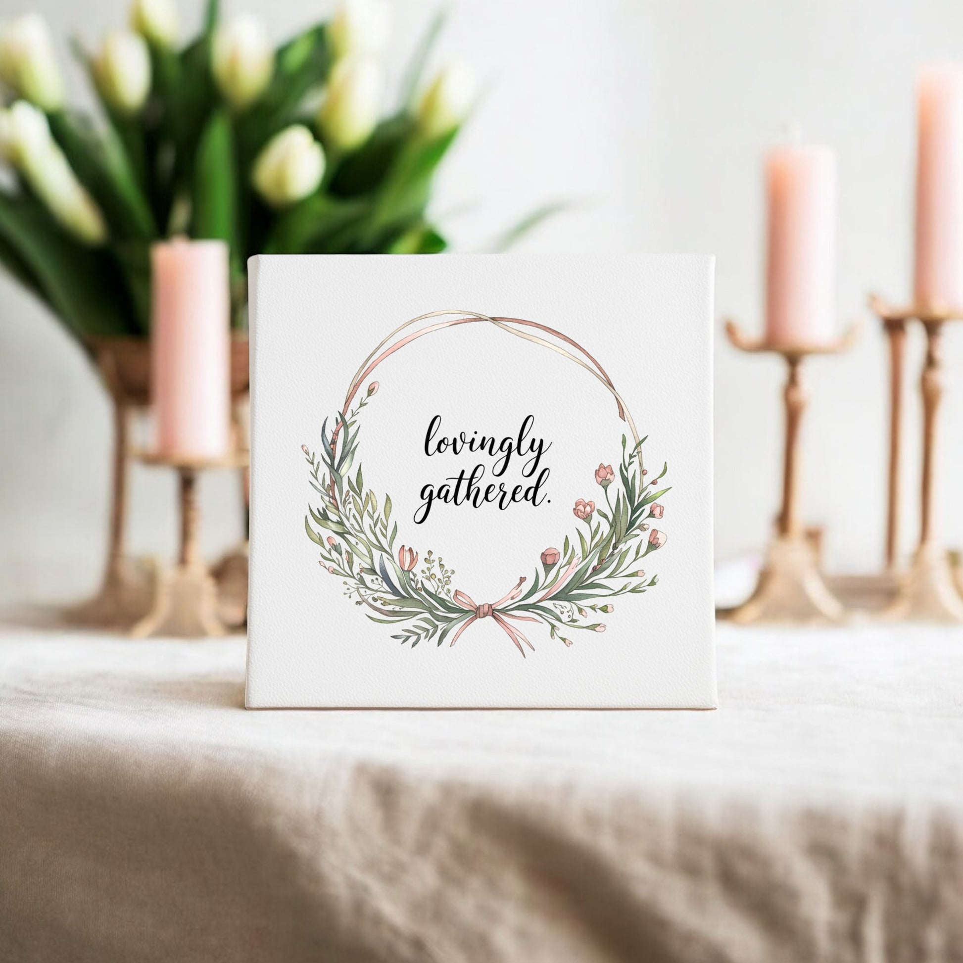 The Mirabilia Boutique Lovingly Gathered Canvas – 8x8 Botanical Wreath Wall Art sits on a beige tablecloth, with white tulips in a vase and three lit pink candles—creating a soft, elegant gathering home decor setting.