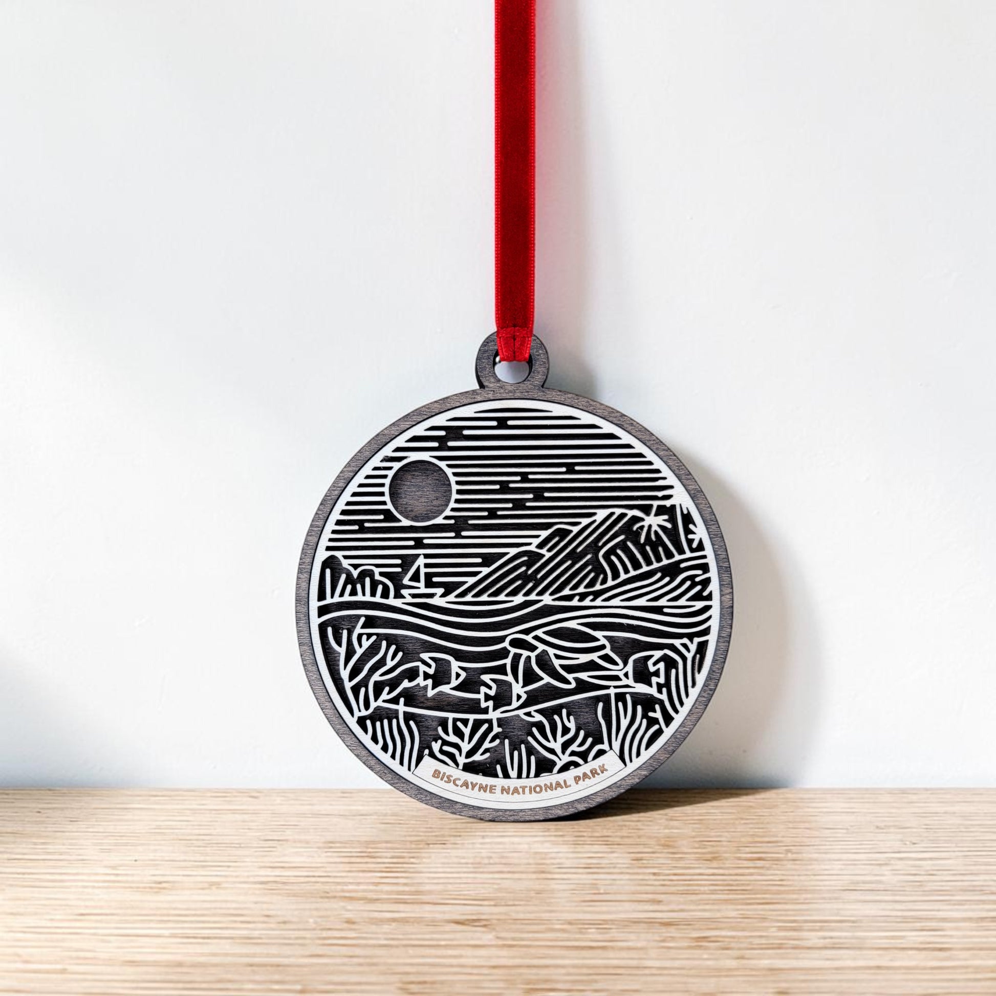 The Biscayne National Park Line Art Ornament by Mirabilia Boutique is a round, wooden keepsake featuring a black and white landscape design with mountains, water, and a sun or moon. It hangs from a red ribbon and displays "Biscayne National Park" at the bottom.