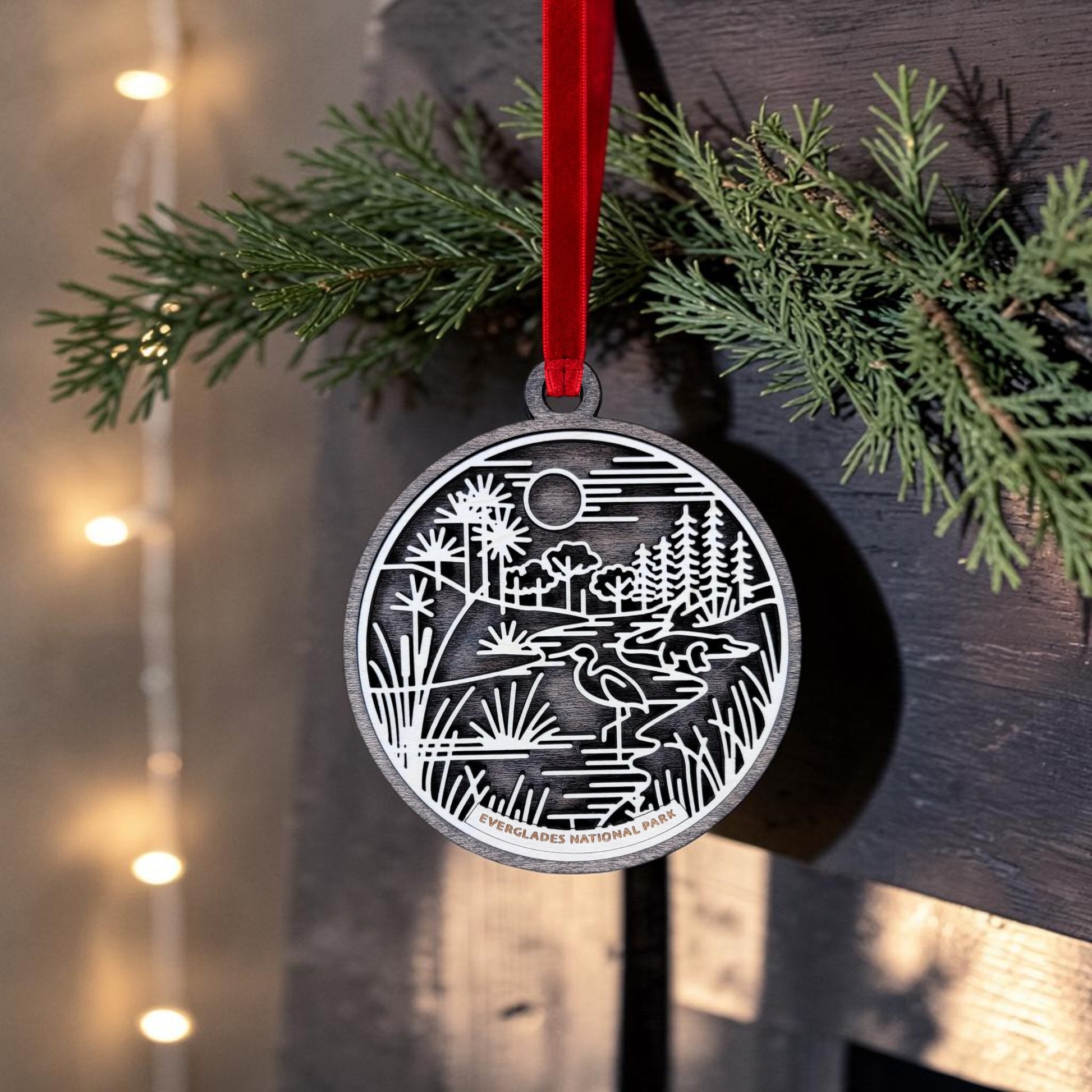 A round metallic Everglades National Park Line Art Christmas Ornament by Mirabilia Boutique, featuring an engraved park scene, hangs from a red ribbon on a pine branch with warm string lights glowing in the background.