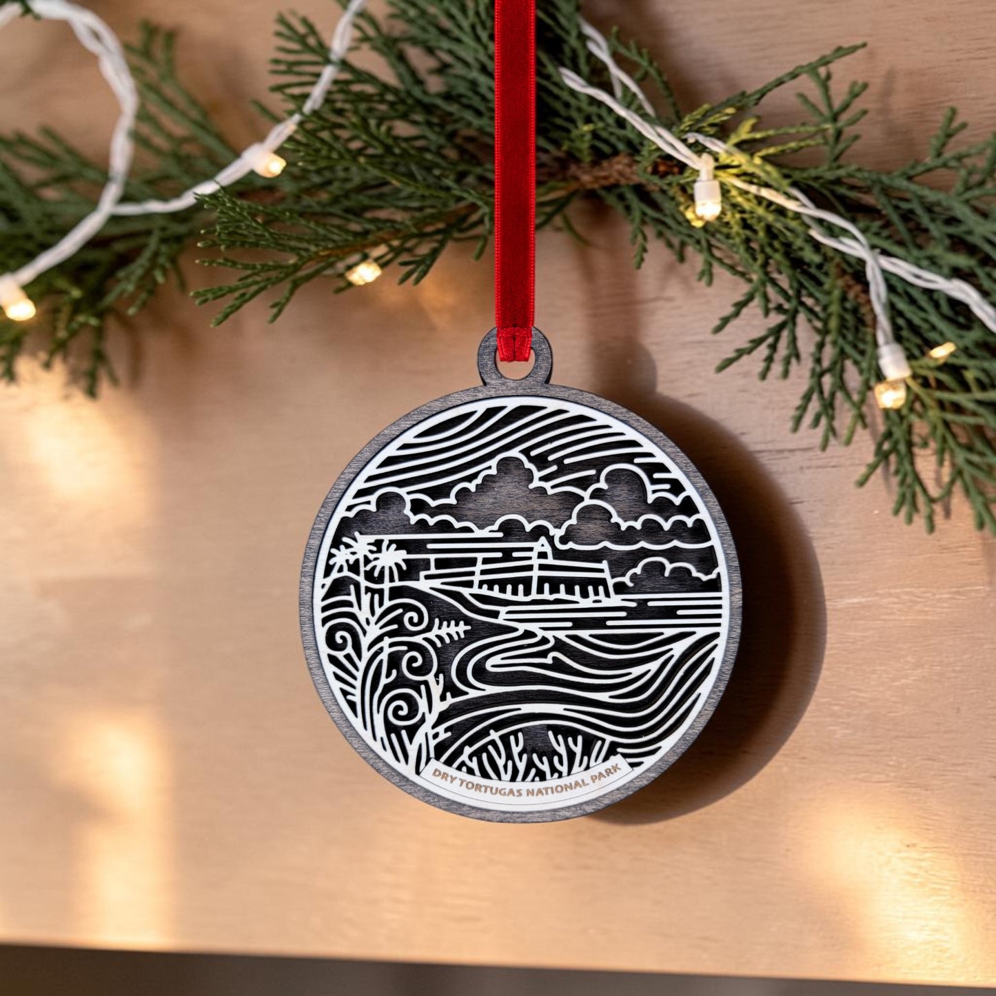 The Dry Tortugas National Park Christmas Ornament by Mirabilia Boutique features a black and white scenic design inspired by Dry Tortugas National Park and Fort Jefferson, complete with a red ribbon and the text “Dry Tortugas National Park.” This oval ornament is displayed hanging on a wooden surface with pine garland and string lights in the background.