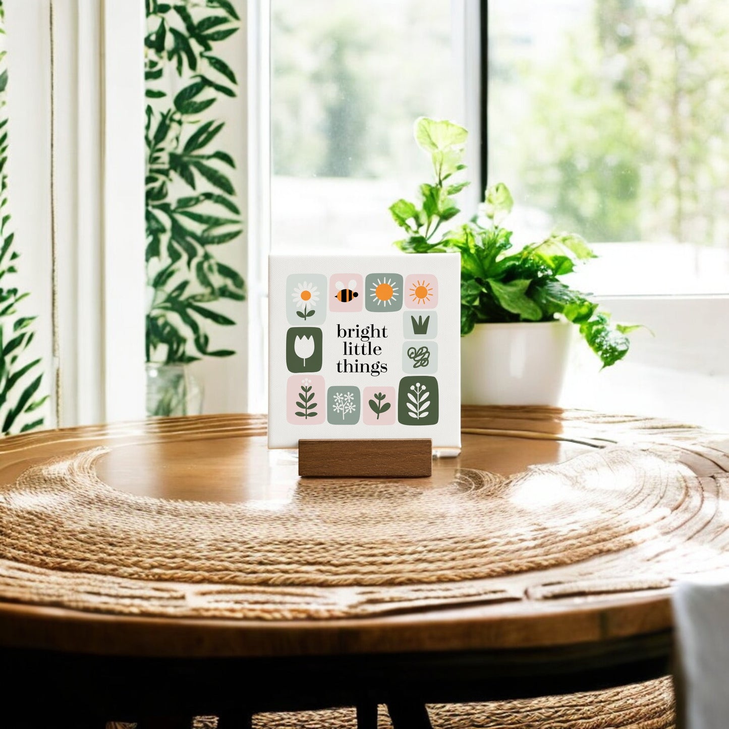 The Bright Little Things Mini Canvas Sign by Mirabilia Boutique features floral illustrations and uplifting text, displayed on a wooden table with greenery and sunlight for a charming handcrafted home decor accent.