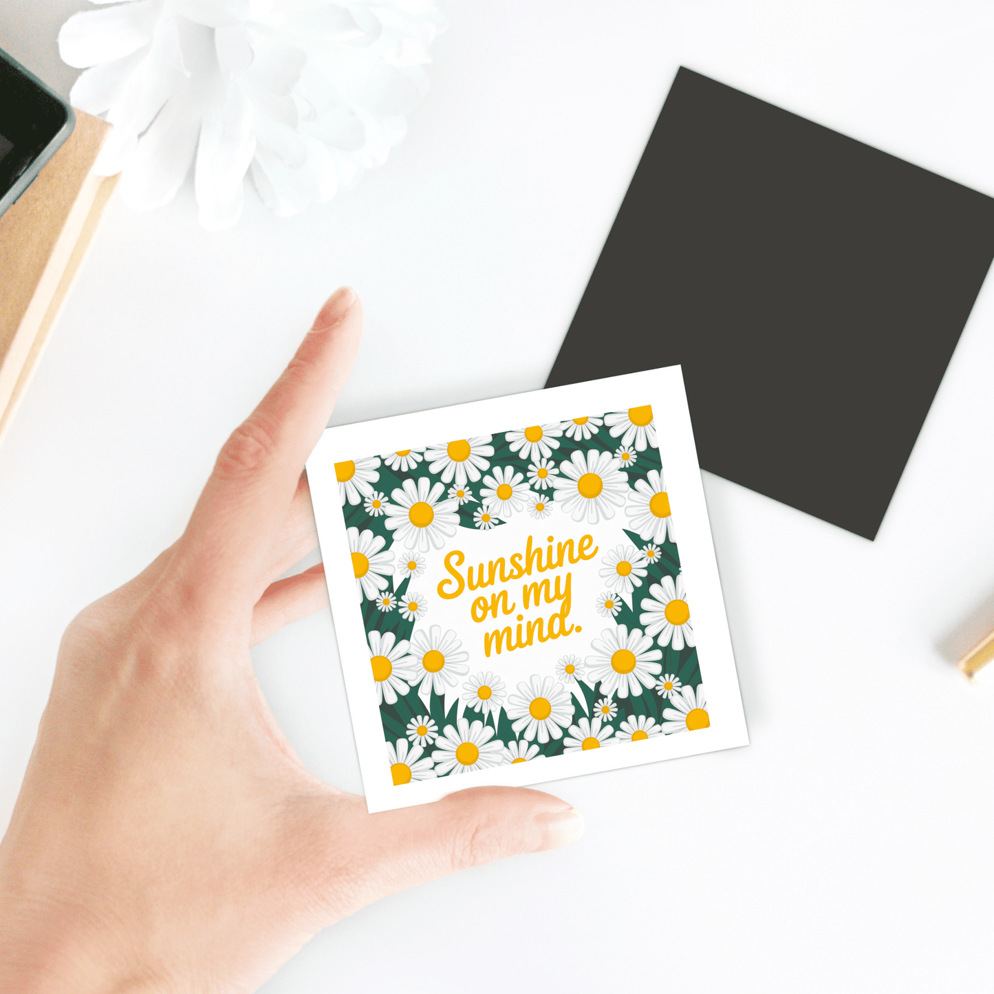 A hand holds the Mirabilia Boutique Sunshine on My Mind Magnet – Daisy Floral Fridge Art, featuring daisies and yellow text. Behind it are a black square, part of a white flower, and objects on a white surface.