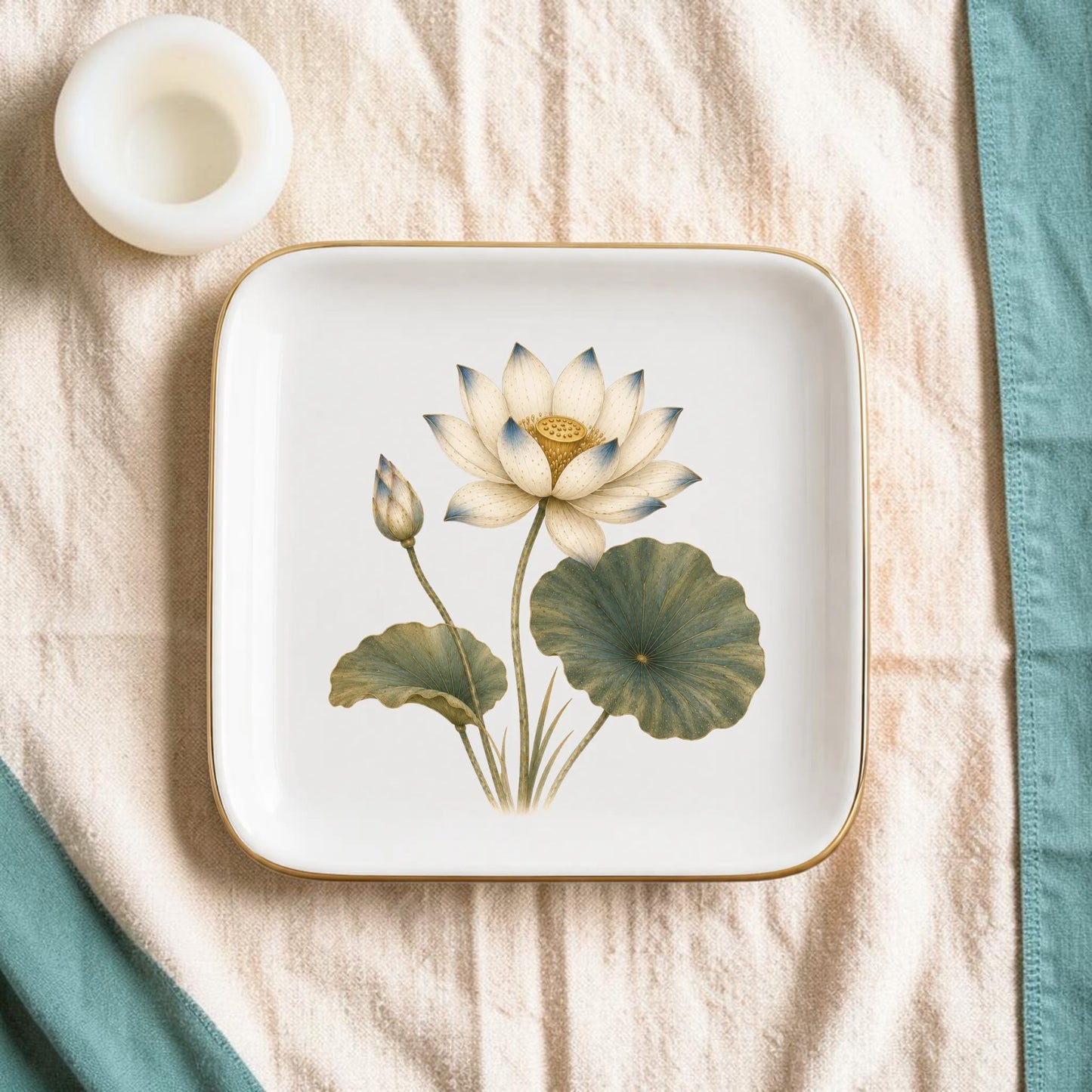 The Flowering Lotus Jewelry Tray | Botanical Catchall by Mirabilia Boutique, featuring a delicate floral design, rests on a beige textured cloth beside a small white round candle and a teal napkin along the edge.