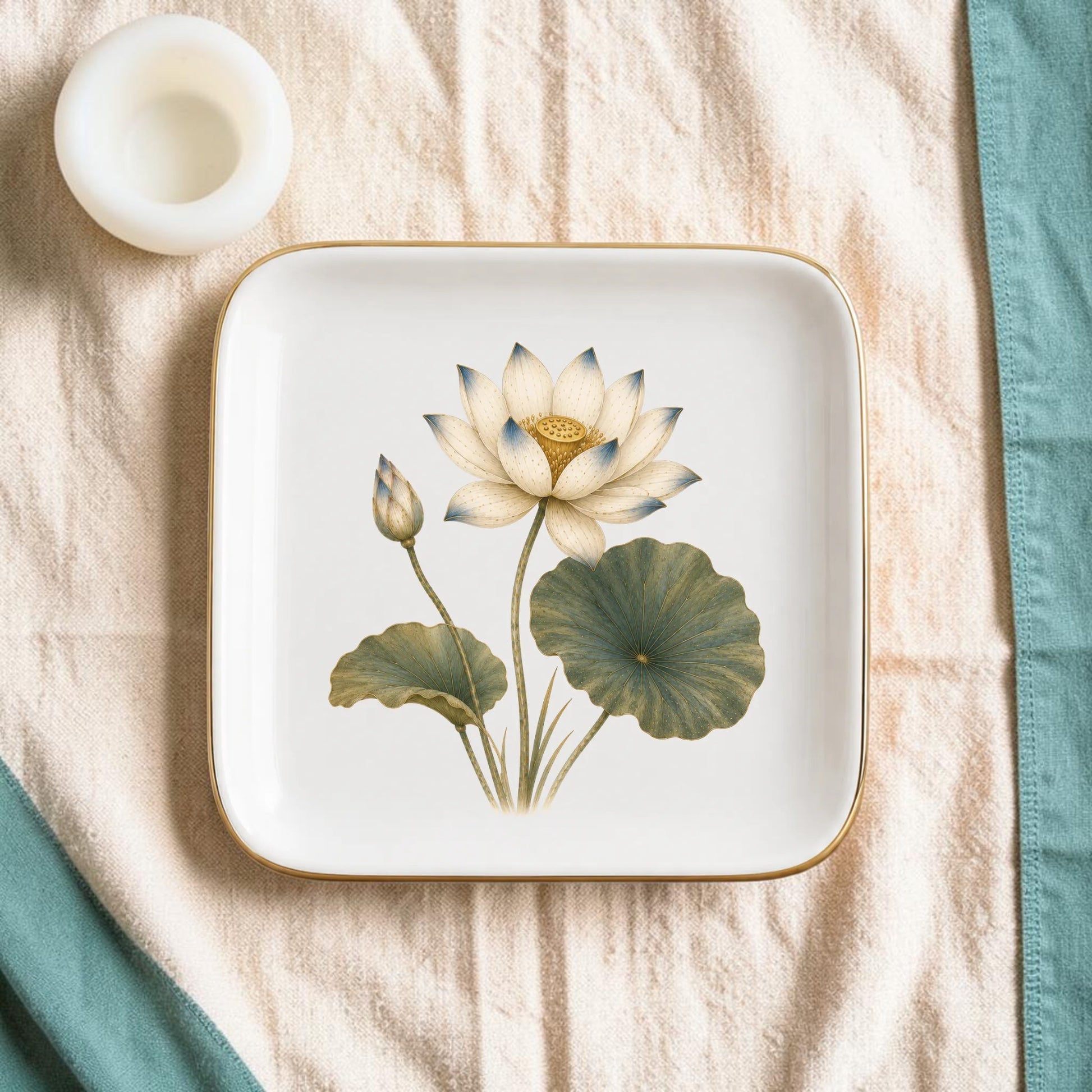 The Flowering Lotus Jewelry Tray | Botanical Catchall by Mirabilia Boutique, featuring a delicate floral design, rests on a beige textured cloth beside a small white round candle and a teal napkin along the edge.
