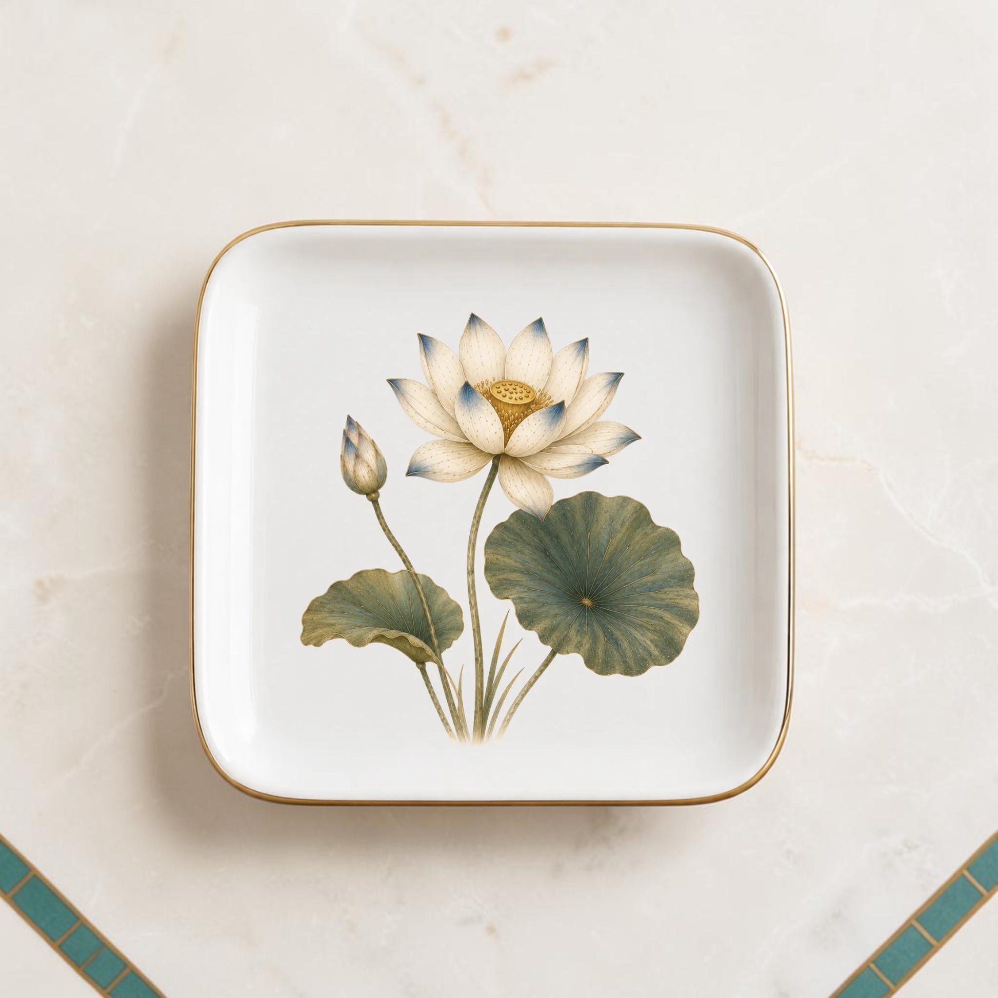 Mirabilia Boutique’s Flowering Lotus Jewelry Tray | Botanical Catchall is a square ceramic tray with gold trim, showcasing a detailed illustration of a white lotus flower, a lotus bud, and two green leaves set against a marble background with a tiled corner.