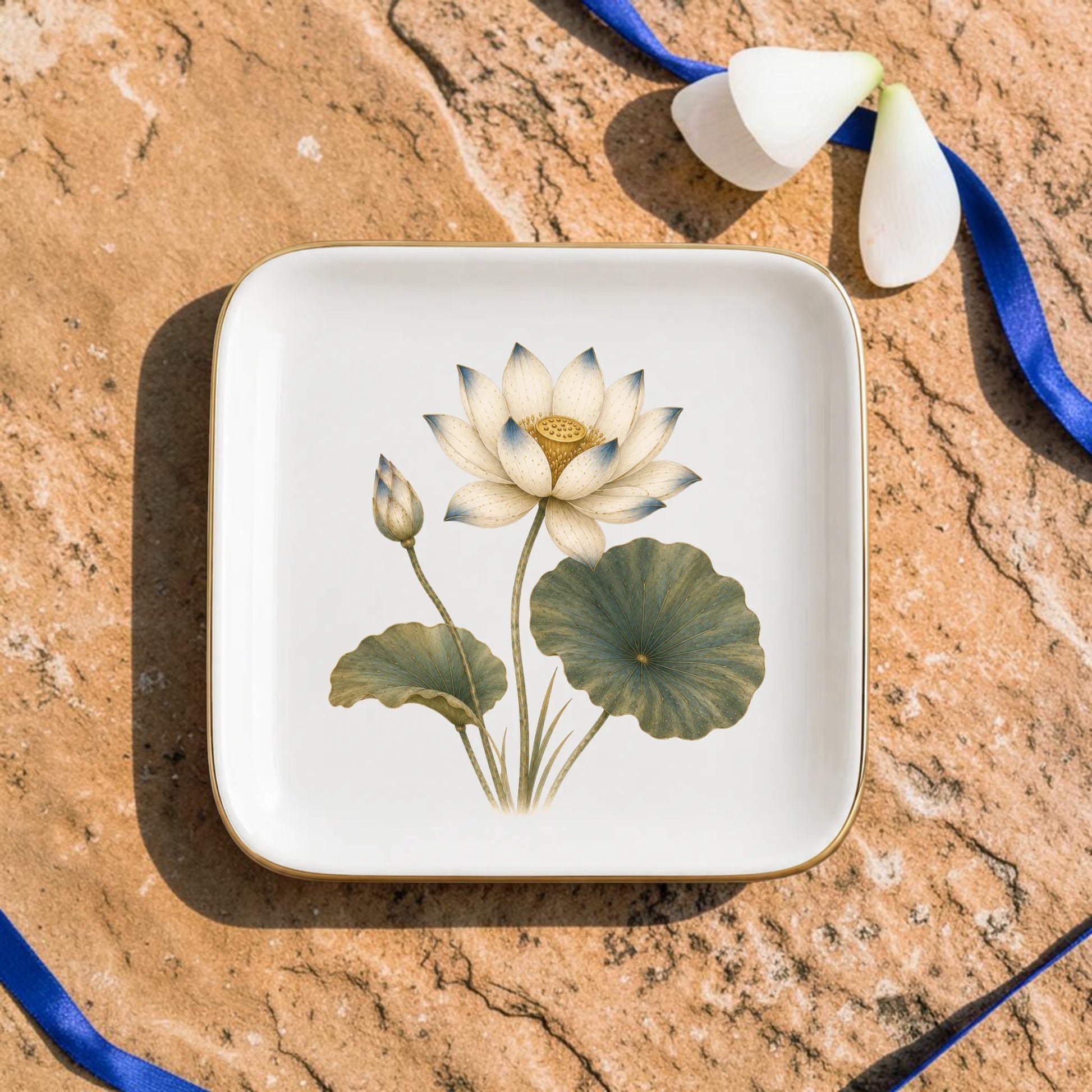 The Flowering Lotus Jewelry Tray | Botanical Catchall by Mirabilia Boutique, a square ceramic decorative piece featuring a lotus flower illustration, sits on a stone surface adorned with blue ribbon and scattered white flower petals.
