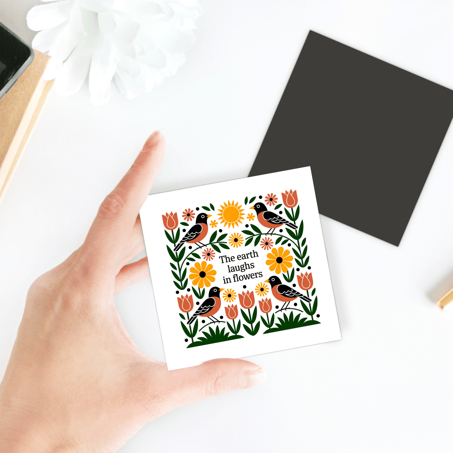 A hand displays Mirabilia Boutique's "The Earth Laughs in Flowers Floral Magnet," featuring vibrant flowers, birds, and leaves with the quote: "The earth laughs in flowers." A black square and white flower rest nearby on a white surface.