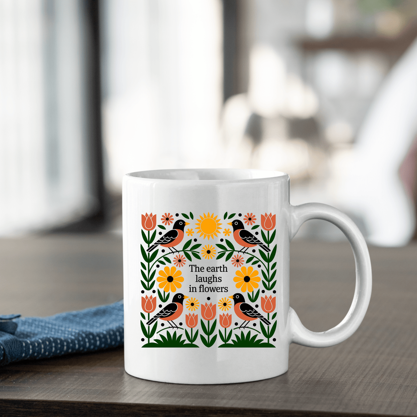 The Earth Laughs in Flowers Spring Mug by Mirabilia Boutique features colorful floral and bird designs with the quote "The earth laughs in flowers." Ideal for nature lovers, it sits beside a folded blue cloth on a softly blurred table.