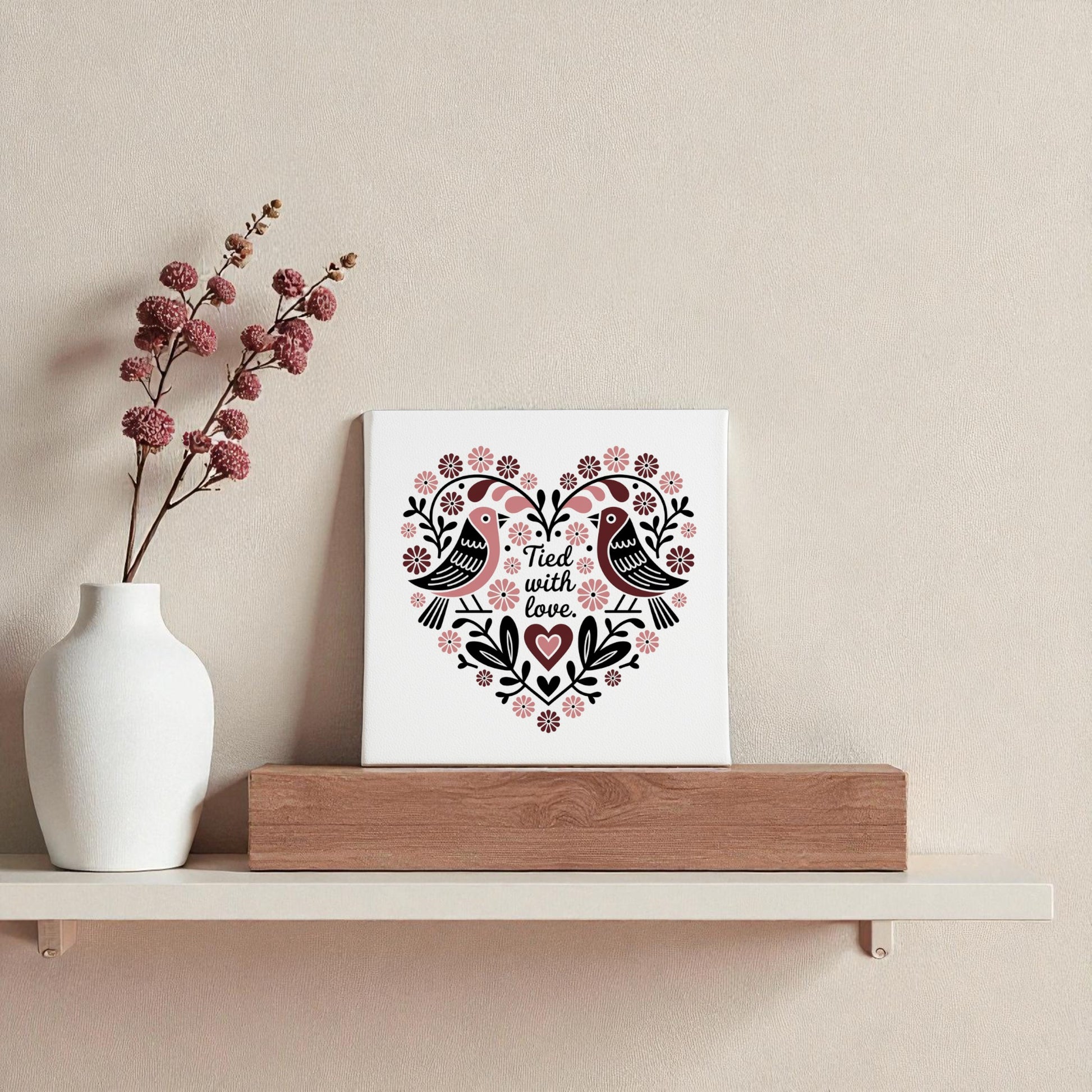 A white vase with pink berry branches and the Mirabilia Boutique "Tied With Love Folk Art Valentine Canvas," featuring Scandinavian-inspired lovebirds, are displayed on a minimalist shelf against a light beige wall.