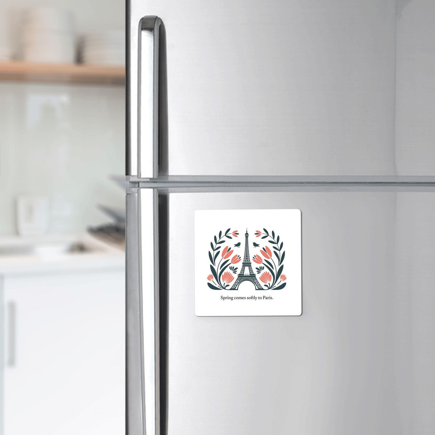 A silver fridge displays the Mirabilia Boutique “Spring Comes Softly to Paris – Floral Eiffel Magnet,” featuring the Eiffel Tower, floral accents, and text. Blurred kitchen counters and shelves appear in the background.