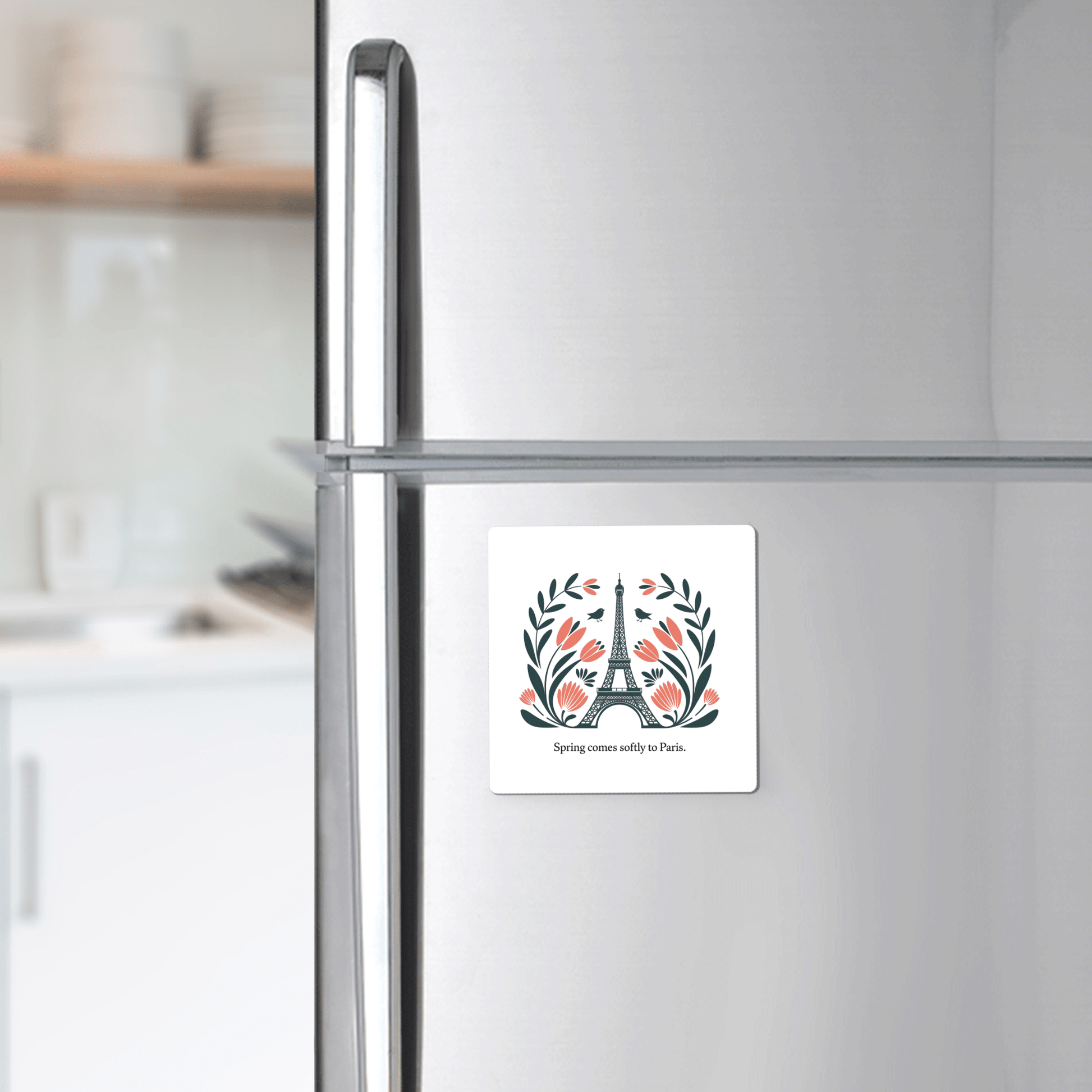 A silver fridge displays the Mirabilia Boutique “Spring Comes Softly to Paris – Floral Eiffel Magnet,” featuring the Eiffel Tower, floral accents, and text. Blurred kitchen counters and shelves appear in the background.