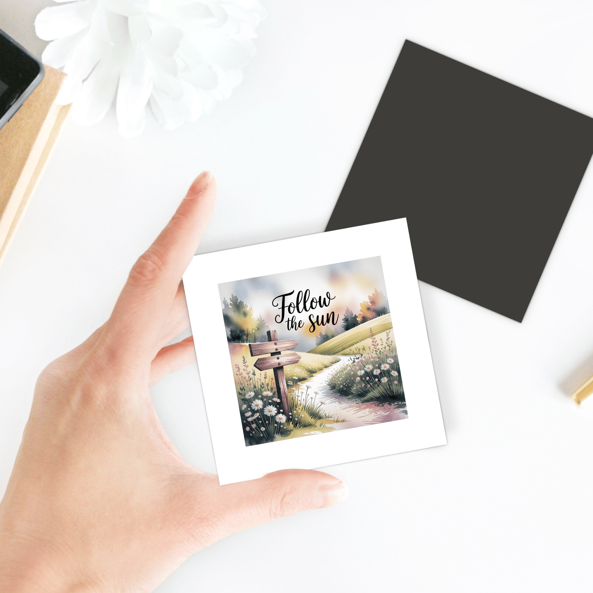 A hand holds the Mirabilia Boutique "Follow the Sun Magnet"—a meadowcore decor piece for fridge or locker, featuring a path, flowers, and uplifting quote. Perfect to brighten your space with nature-inspired style.