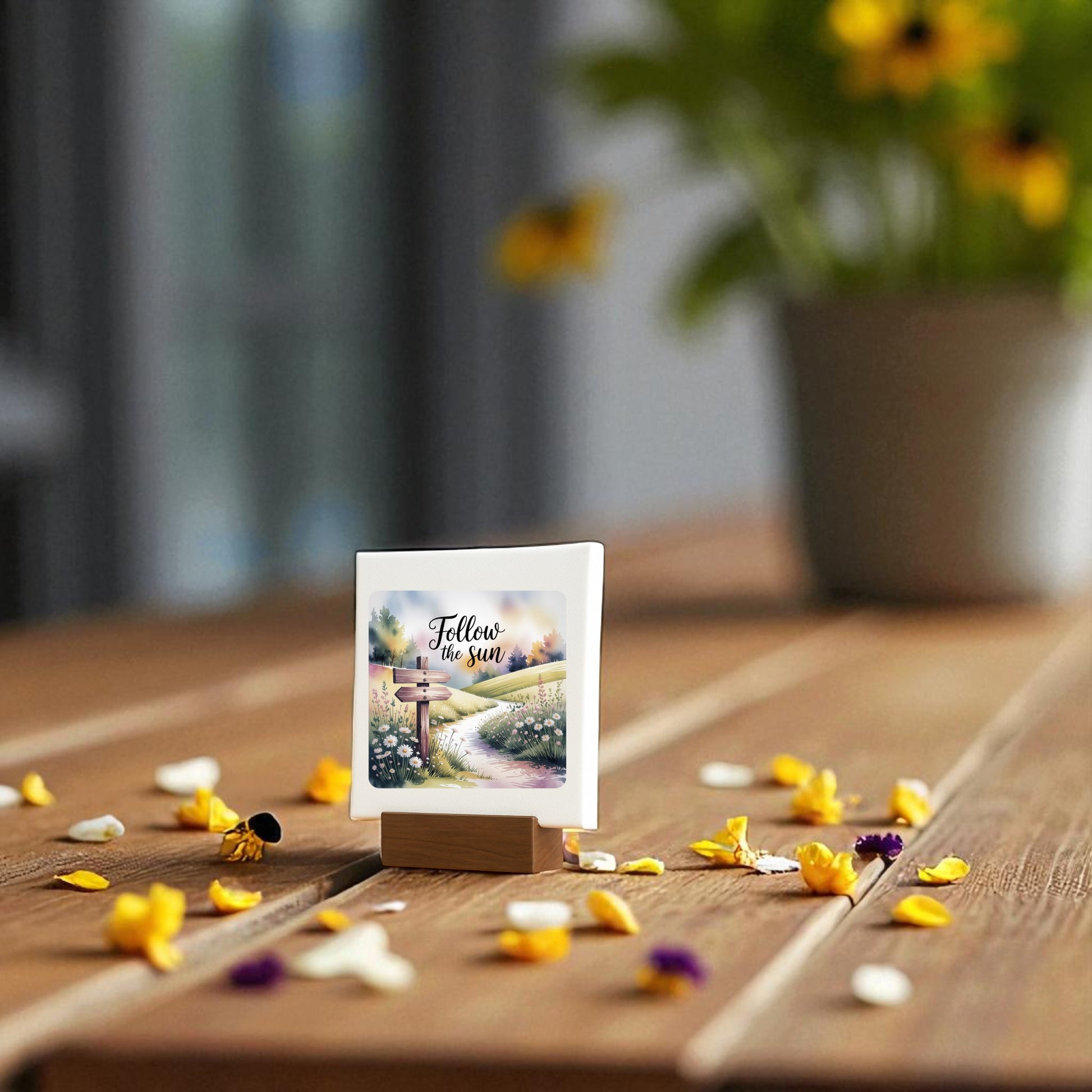 The Follow the Sun Mini Sign by Mirabilia Boutique, perfect as inspirational nature decor, sits on a wooden table with flower petals; a yellow-flowered potted plant is softly blurred in the background.