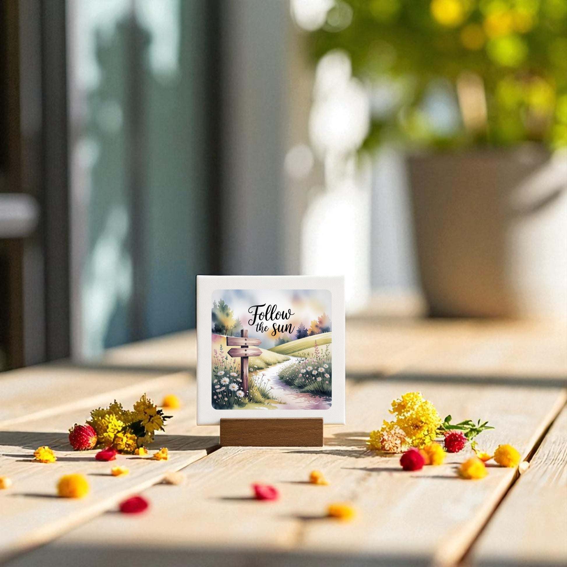 The Follow the Sun Mini Sign by Mirabilia Boutique rests on a wooden table with yellow and red flowers, while a blurred potted plant and sunlight in the background create an inviting, inspirational nature decor vibe.