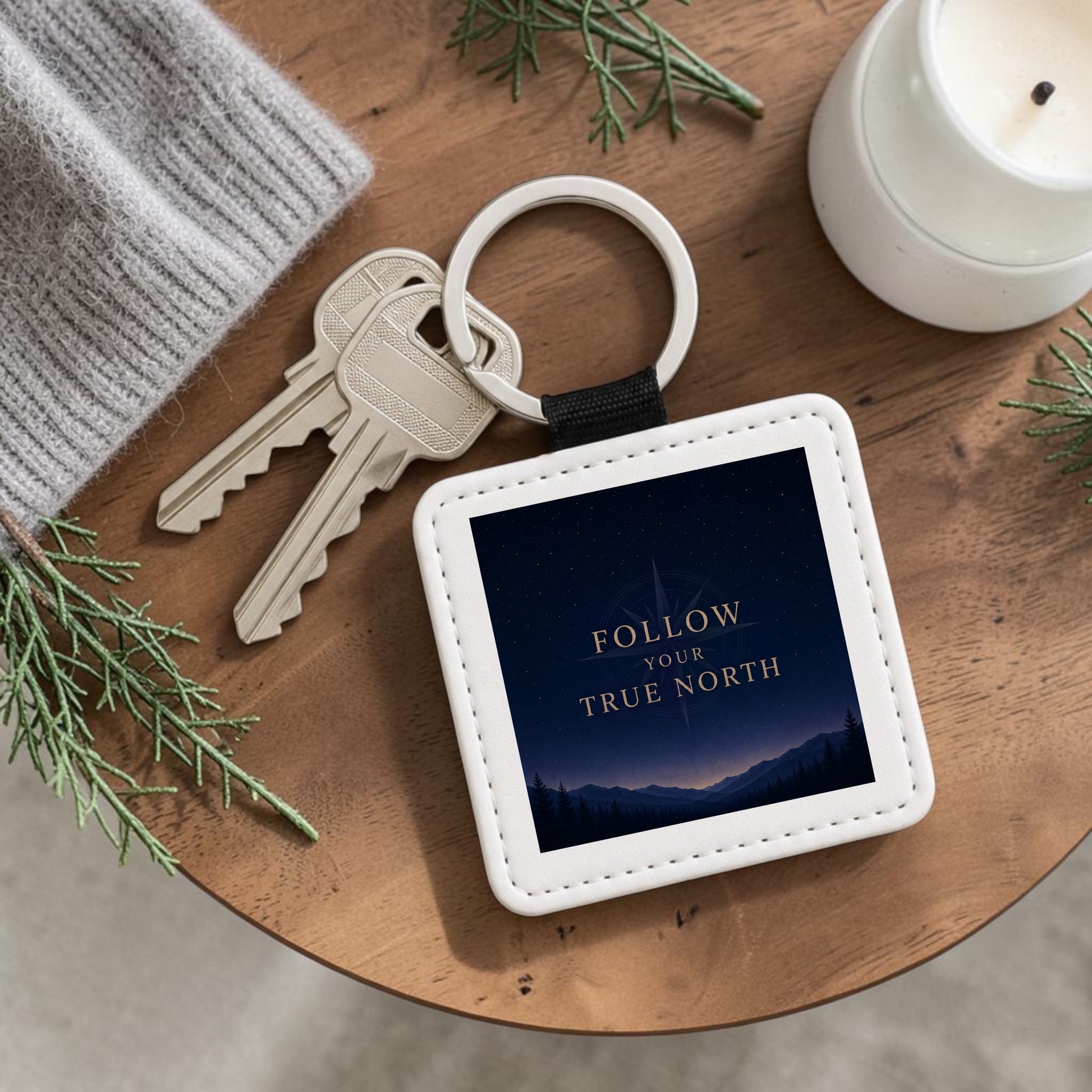 The Follow Your True North Celestial Keychain by Mirabilia Boutique, featuring a square design and the inspiring phrase “Follow Your True North,” rests on a wooden table beside two keys, a gray knit accessory, lush greenery, and a white candle—capturing the charm of celestial navigation and personal discovery.