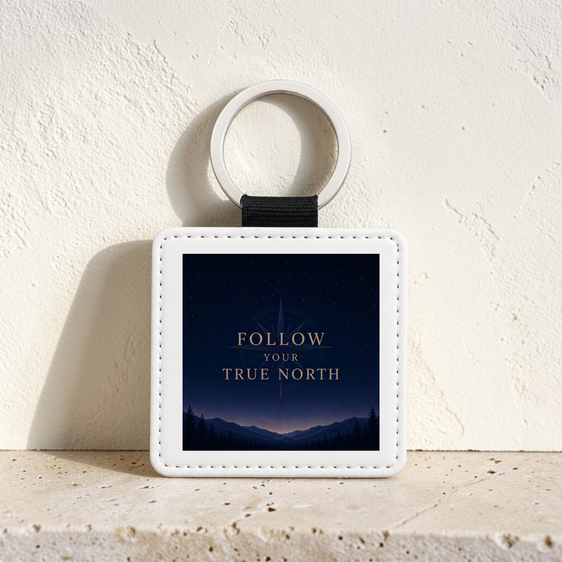 The Follow Your True North Celestial Keychain by Mirabilia Boutique features a white, square design showcasing a night sky, mountains, and trees, with “Follow Your True North” written in gold letters—capturing the spirit of celestial navigation.