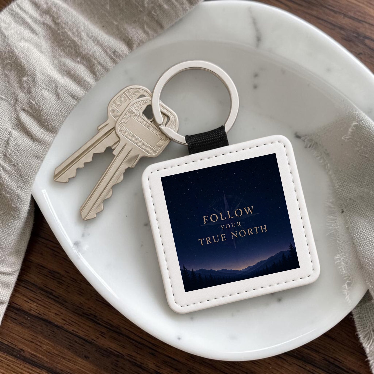 A square Follow Your True North Celestial Keychain by Mirabilia Boutique, adorned with keys, rests on a white dish. The keychain features a night sky scene with mountains and the inspiring phrase “FOLLOW YOUR TRUE NORTH” in gold letters, capturing the essence of celestial navigation. A light-colored fabric is placed nearby.