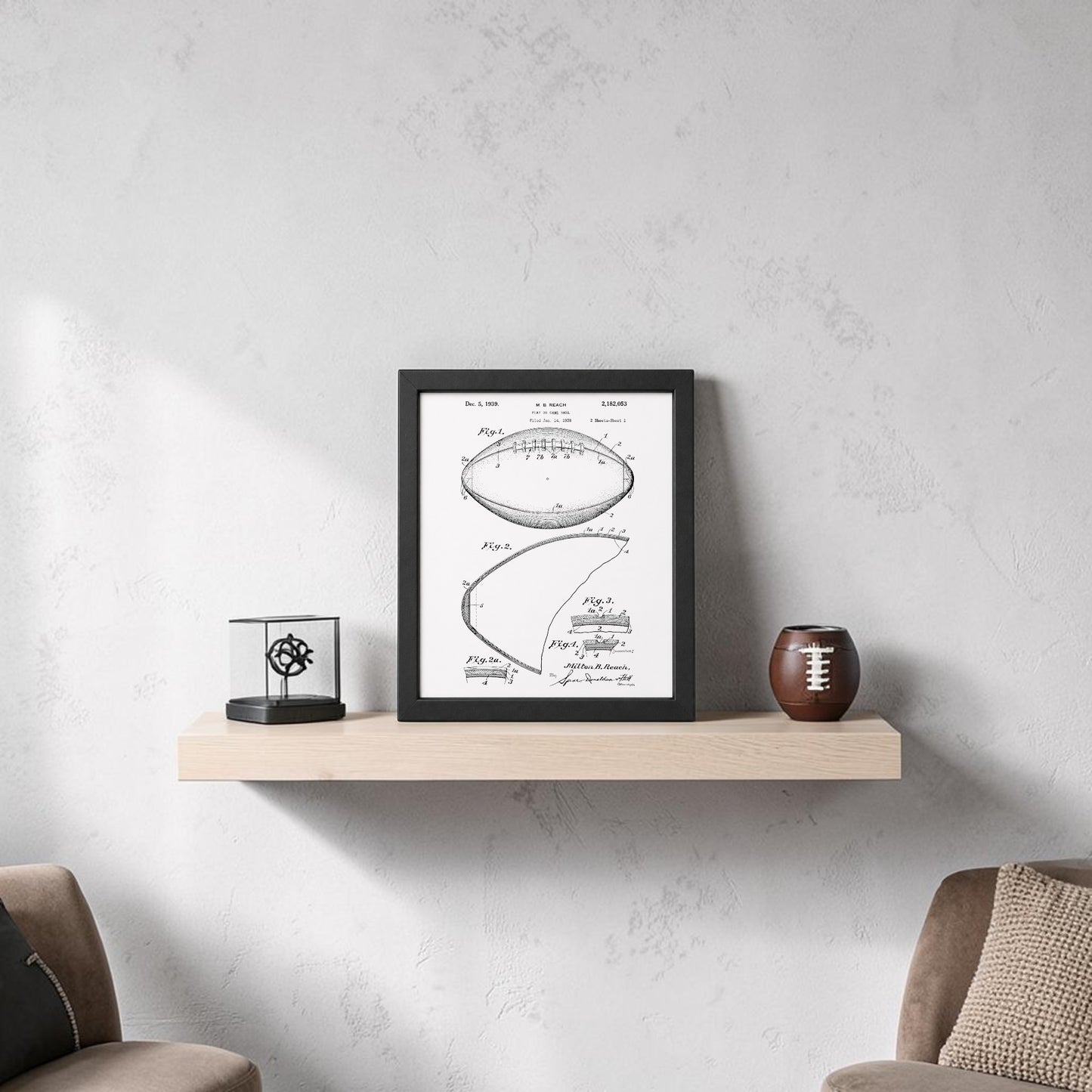 A minimalist room includes a light wood floating shelf with the Mirabilia Boutique American Football Patent Art Print 8x10, a small glass display case, and a decorative football. Neutral-toned chairs and a textured light gray wall complete the look.