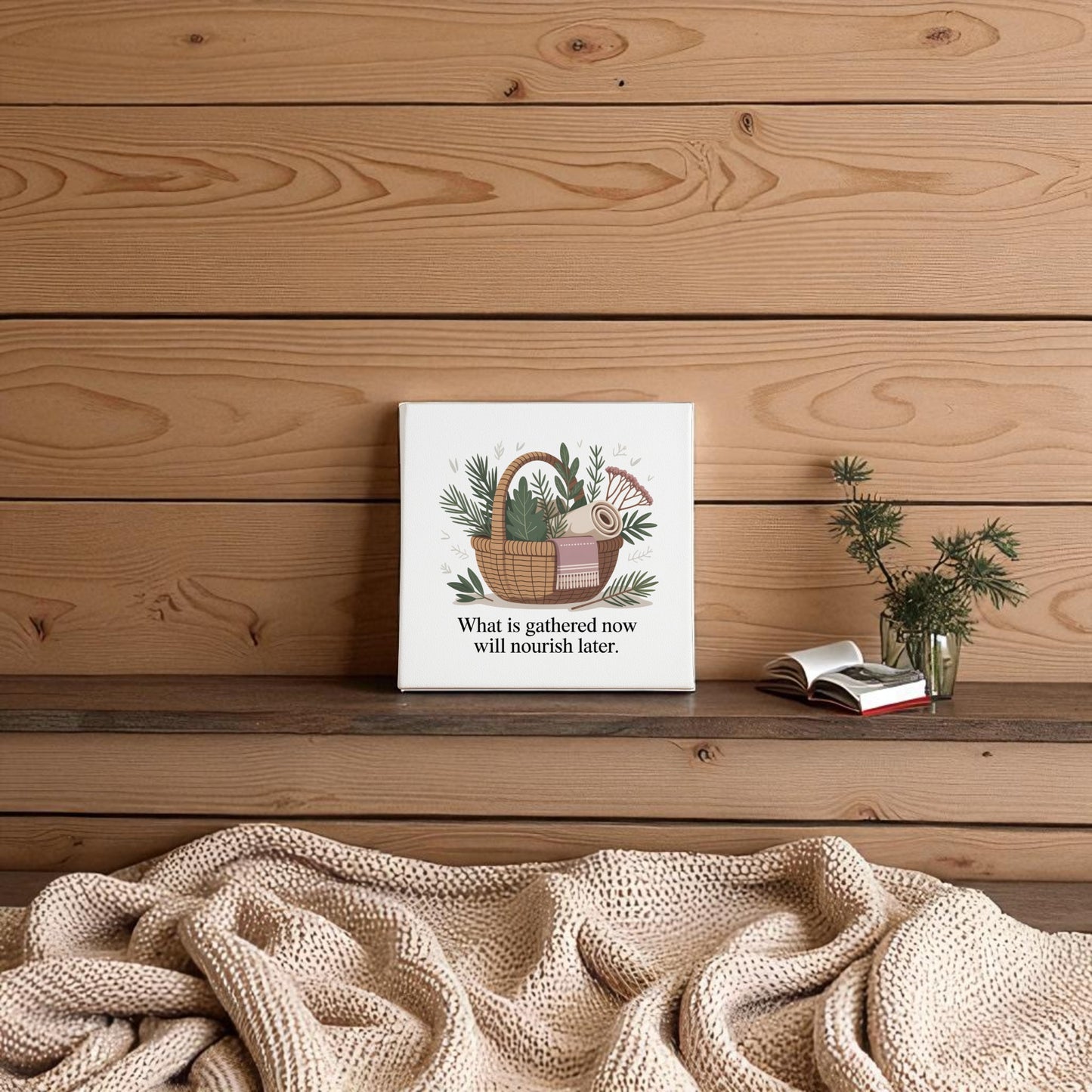 A cozy scene with a textured blanket on a wooden bench, a plant in glass, an open book, and the Mirabilia Boutique “Herbal Basket Canvas Art | What Is Gathered Quote Print” against a wood-paneled wall.