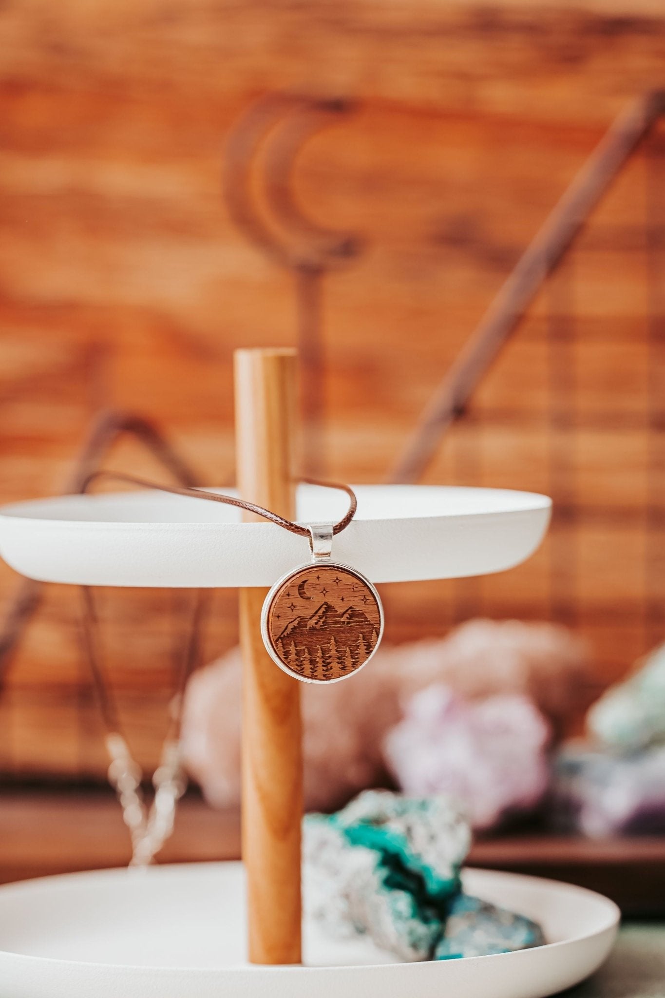 The Engraved Wood Pendant Necklace – Mountain Night Sky Design by Mirabilia Boutique hangs on a brown cord from a wooden stand with white trays; blurred crystals and wood background create a boho display.