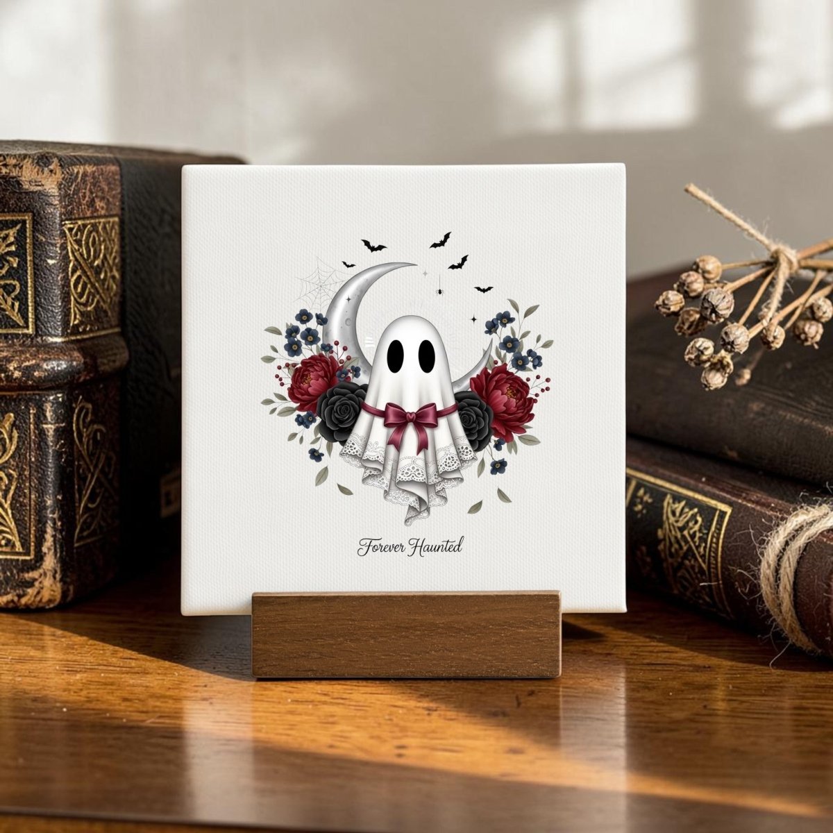 Introducing the Forever Haunted Mini Canvas Sign by Mirabilia Boutique—a charming, small square artwork featuring an adorable ghost adorned with red roses, surrounded by a crescent moon, black bats, and delicate lace. Resting on a wooden surface beside vintage books, this piece makes the perfect gothic cottagecore decor or Halloween home accent. The haunted mini canvas sign is finished with the phrase "Forever Haunted" below the ghost for an extra touch of spooky elegance.