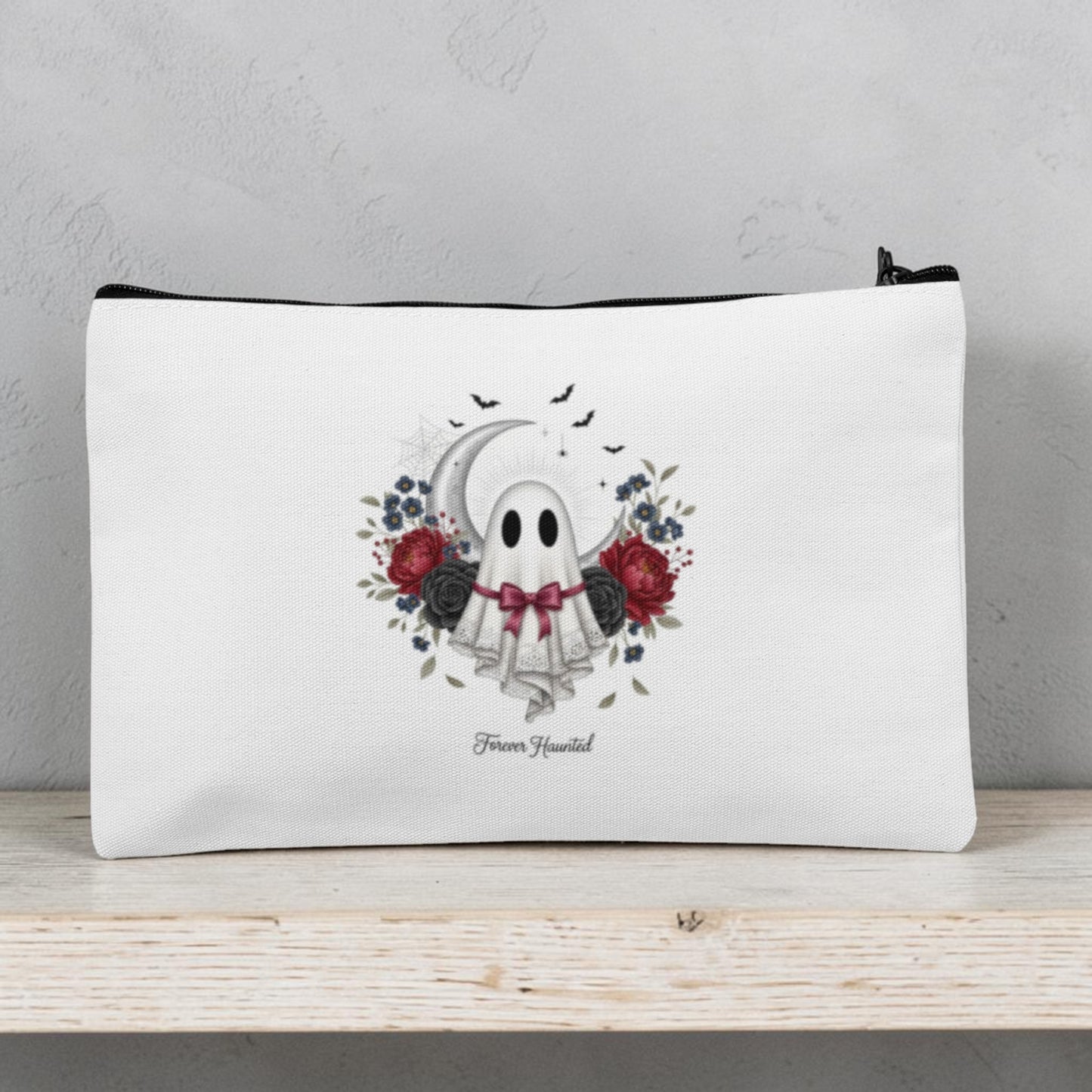 The Forever Haunted Makeup Bag | Gothic Ghost Pouch by Mirabilia Boutique features a white zippered design decorated with an adorable ghost wearing a floral crown and bowtie, surrounded by red and black roses, leaves, and bats. The phrase “Boo Yourself” appears beneath the ghost. Perfect as a spooky season accessory or Halloween makeup bag, this gothic cosmetic pouch is displayed on a light wooden shelf.