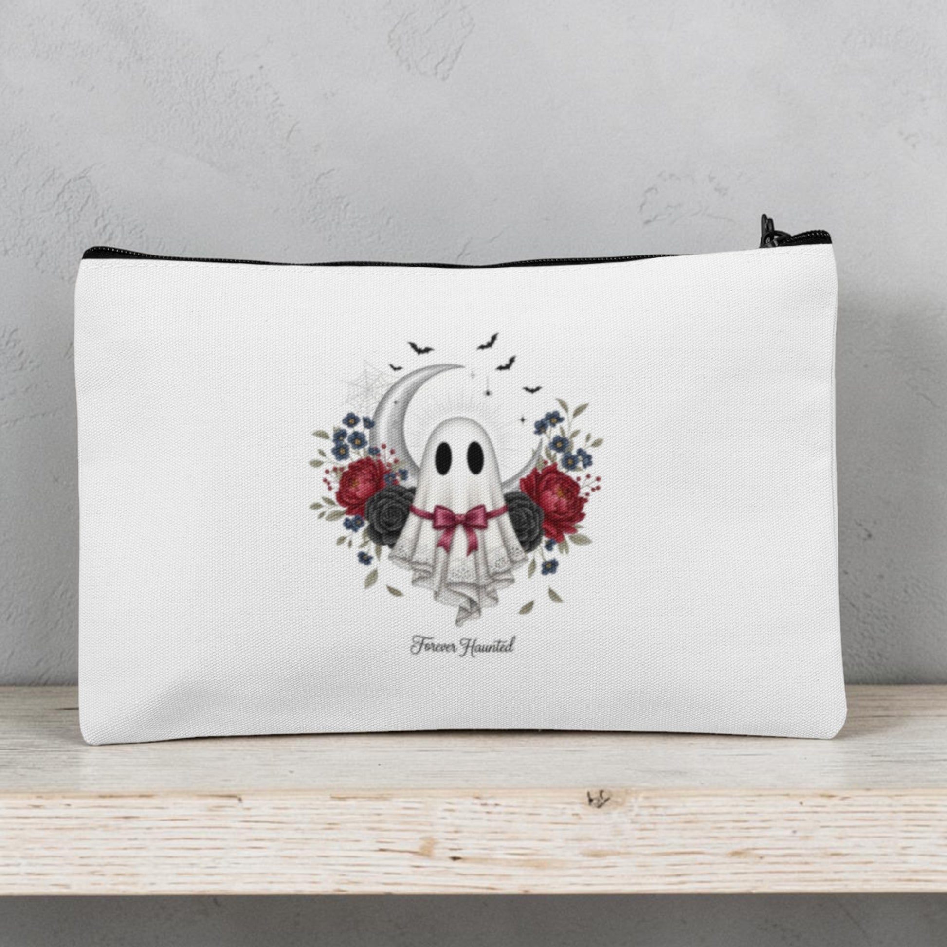 The Forever Haunted Makeup Bag | Gothic Ghost Pouch by Mirabilia Boutique features a white zippered design decorated with an adorable ghost wearing a floral crown and bowtie, surrounded by red and black roses, leaves, and bats. The phrase “Boo Yourself” appears beneath the ghost. Perfect as a spooky season accessory or Halloween makeup bag, this gothic cosmetic pouch is displayed on a light wooden shelf.