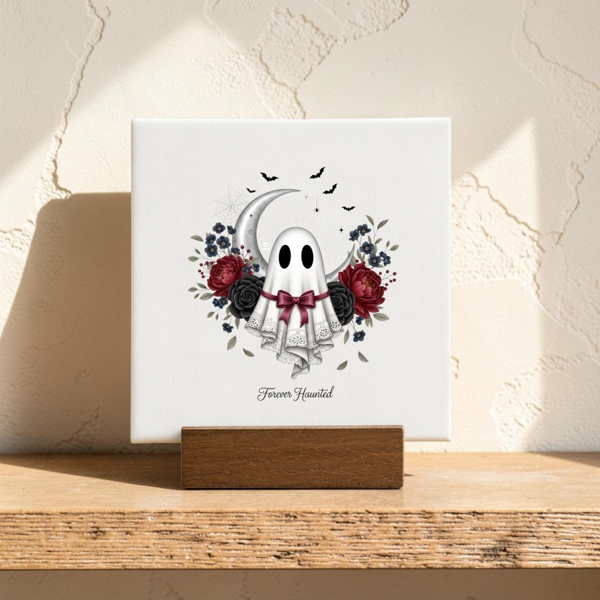 The Forever Haunted Mini Canvas Sign by Mirabilia Boutique features a charming ghost with a red bow, surrounded by roses, a crescent moon, bats, and delicate lace. The artwork is accented with the phrase "Forever Haunted" beneath the illustration. Perfect for Halloween home decor or gothic cottagecore themes, this mini canvas comes displayed on a wooden stand against a textured wall.