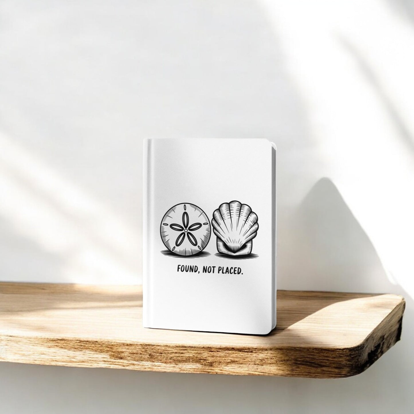 The Found, Not Placed Journal – Coastal Quote Notebook by Mirabilia Boutique, featuring white seashell designs and the phrase "Found, not placed.", rests on a sunlit wooden shelf casting soft shadows against a white wall.