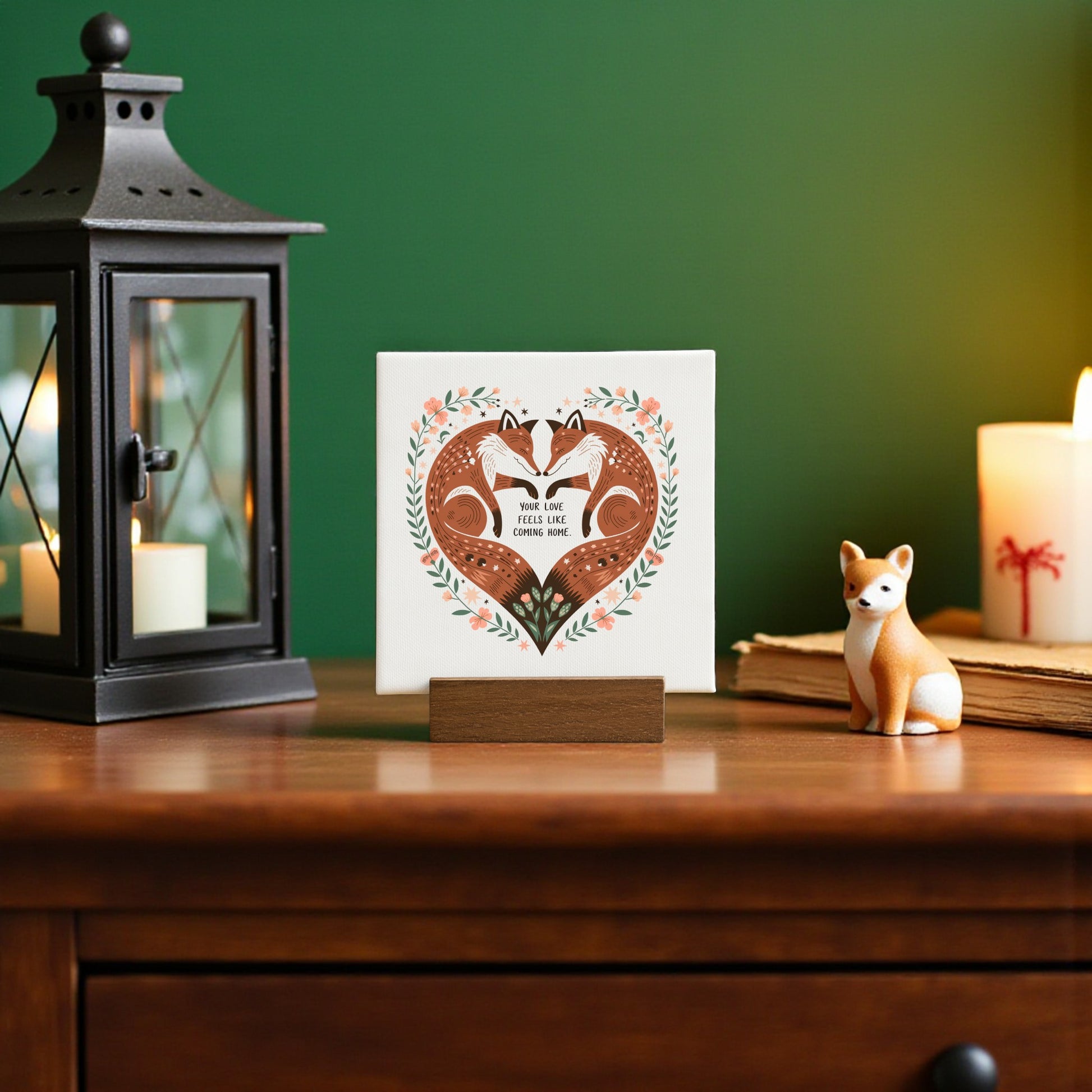 A wooden table displays a lantern, candle, fox figurine, and the Mirabilia Boutique Fox Love Mini Canvas featuring “Your Love Feels Like Home”—a charming rustic woodland decor piece celebrating love.