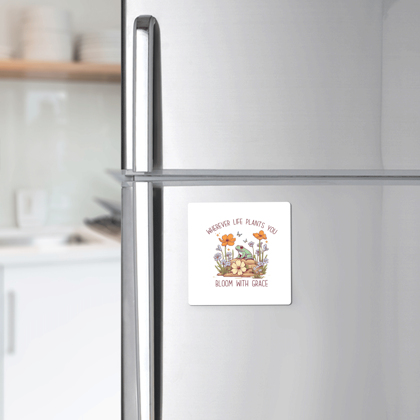 A Mirabilia Boutique Frog Magnet reads "Bloom With Grace" and features colorful flowers and mushrooms on a stainless steel fridge—an inspirational decor piece from the Frog Magnet | Bloom With Grace Inspirational Gift line. A blurred kitchen appears in the background.