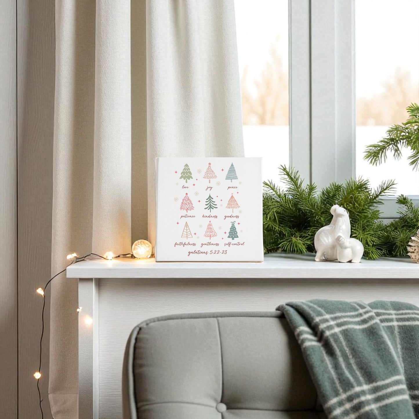 A white shelf by the window displays Mirabilia Boutique’s "Fruits of the Spirit | Christian Christmas Canvas | Galatians 5:22," festive decor, and string lights; beside it, a gray chair with a green plaid blanket makes a cozy Christian Christmas gift scene.