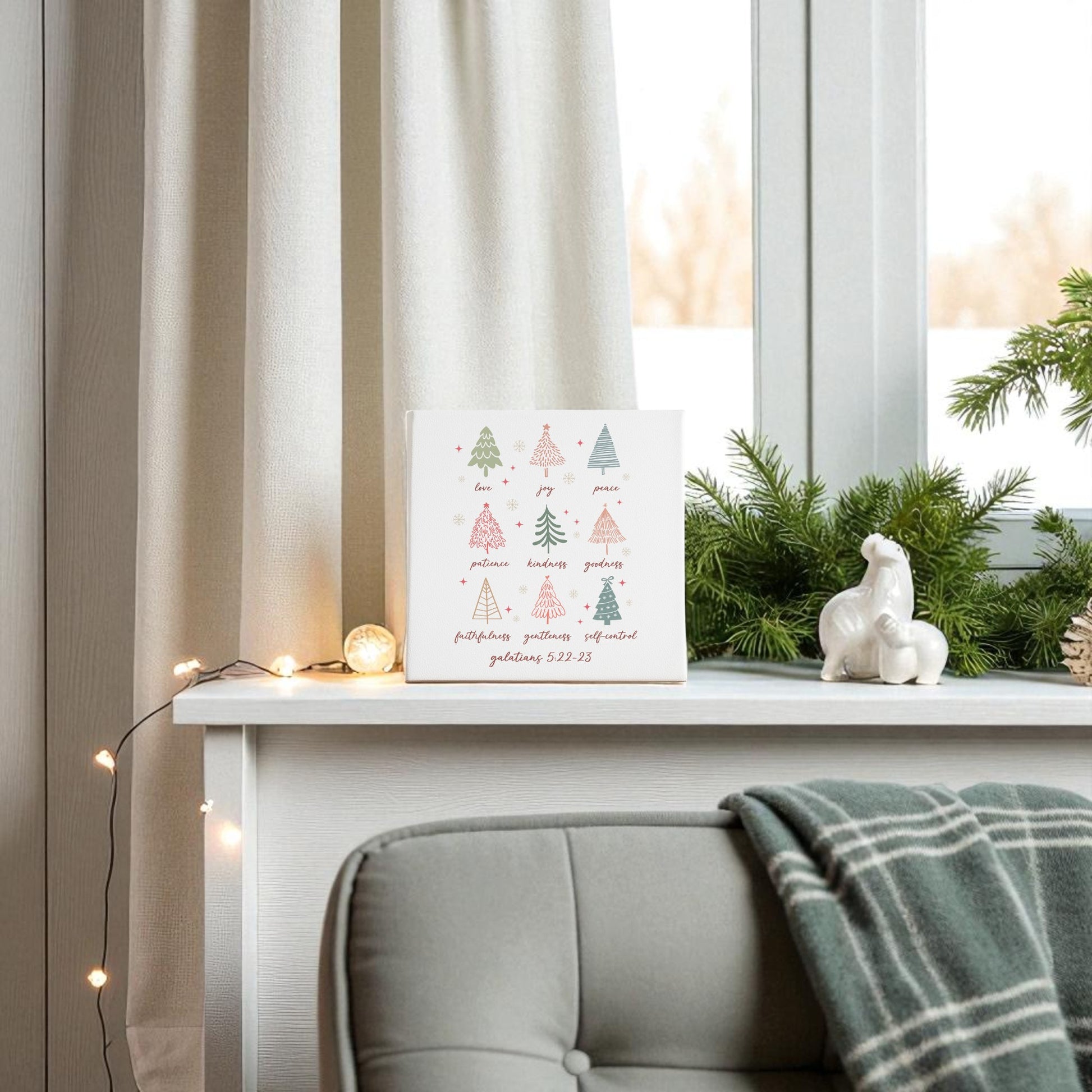 A white shelf by the window displays Mirabilia Boutique’s "Fruits of the Spirit | Christian Christmas Canvas | Galatians 5:22," festive decor, and string lights; beside it, a gray chair with a green plaid blanket makes a cozy Christian Christmas gift scene.